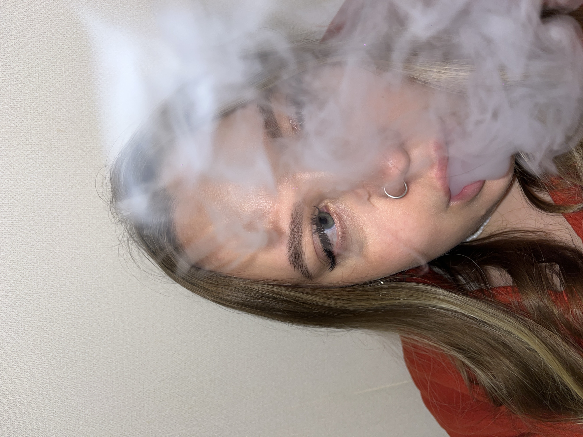 A young woman is partially obscured by a cloud of smoke, likely exhaled from her mouth. She has fair skin, blonde hair with highlights, and a nose ring. Her clothing is a reddish-orange color. The background is a plain, light-colored wall. The image is rotated. There is no verifiable evidence that the image was taken in the city of Krasnodar, Russia.