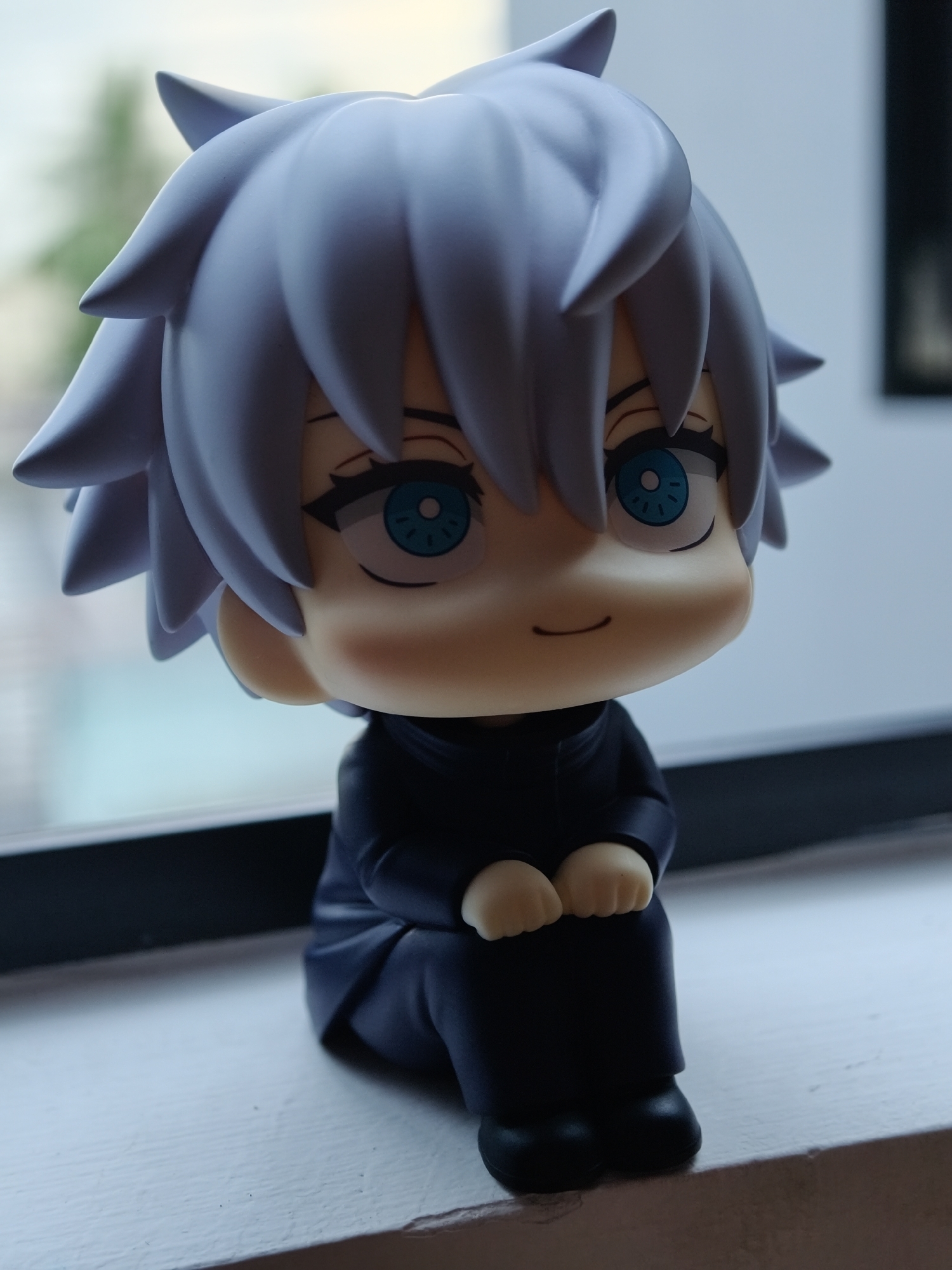 A close-up, vertical, eye-level shot focuses on a small, stylized figurine of an anime character with spiky light blue hair, large bright blue eyes, and rosy cheeks, sitting on a white windowsill. The figurine is dressed in a dark blue outfit, with its hands resting on its lap. The background is blurred, showing a hint of green foliage through a window and a dark rectangular shape, possibly a picture frame. The setting appears to be indoors, near a window with natural light. There is no indication that this scene is located in the city of Da Nang, Vietnam.