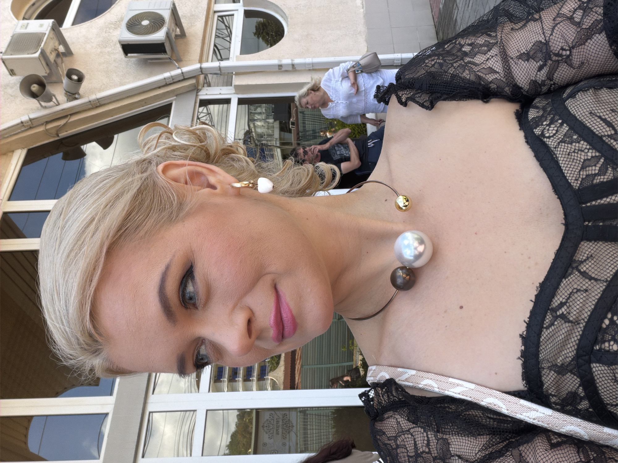 The media file is a portrait of a woman with blonde hair, wearing a black lace top and a striking necklace with large pearl-like beads. She is looking towards the viewer with a slight smile and has striking makeup, including dramatic eyeliner and pink lipstick. Her ear is visible, adorned with a delicate earring.
In the background, other people are visible, including a woman in a white top and a man in a dark t-shirt. The setting appears to be outdoors, in front of a building with air conditioning units mounted on the wall and large windows reflecting the surroundings. The overall impression is that of a casual social gathering or a moment captured during an outing.
The image is oriented vertically, with the subjects' faces angled. The lighting suggests it is daytime, possibly late afternoon, given the soft light and shadows. The background details and architectural style are consistent with a location in Sochi, Russia, as indicated by the context. No explicit text is visible within the frame.