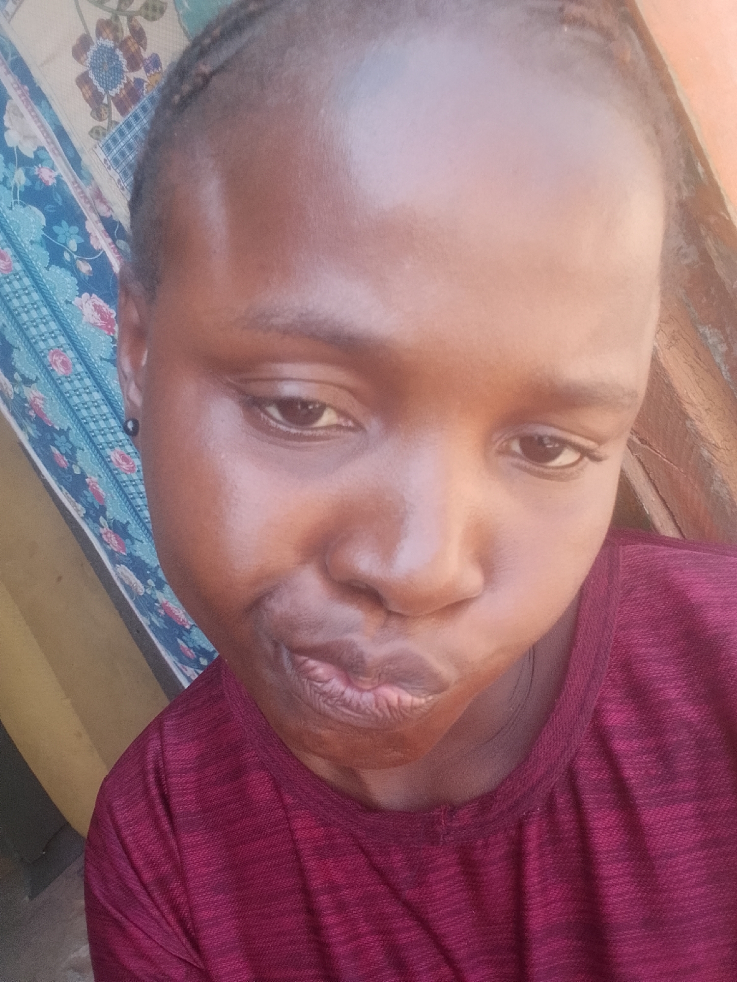 The image is a close-up, slightly angled selfie of a young Black individual. The person has dark skin, short dark hair styled in braids, and is wearing a maroon crew-neck shirt. Their eyes are dark brown, and they have a small black earring in their left ear. The person is looking down and slightly to their left, with their lips pursed. The background is blurred but appears to be indoors, with a patterned fabric on the left and a wooden surface on the right. The lighting suggests it's daytime. There is no discernible information about the location within Pantame, Nigeria.