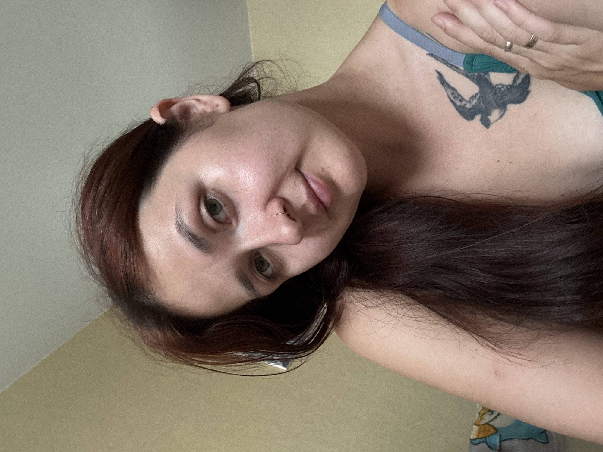 A close-up, rotated image shows a woman with long, dark, reddish-brown hair. She is wearing a blue bra with a grey strap. A tattoo of a bird in flight is visible on her upper chest, near her left shoulder. She has a nose ring and a ring on her right ring finger. The background appears to be a wall and a corner of a room, with a beige wall meeting a lighter colored wall. The lighting suggests it could be daytime, but no specific time can be determined. Her expression is neutral, and the overall mood is relaxed. There is no visible text in the image. The location context of Naberezhnye Chelny, Russia is provided but not discernible from the image itself.