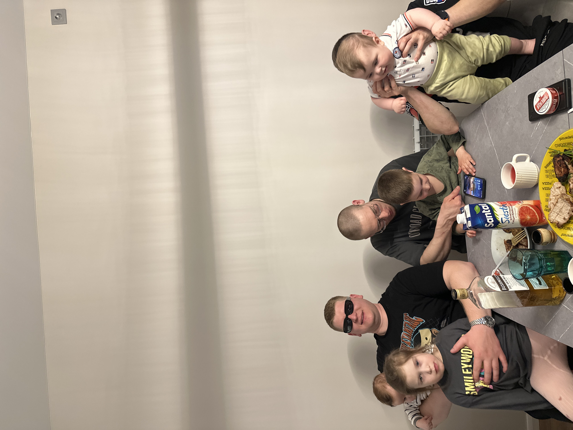 The image shows a group of people gathered around a table, possibly in a home setting. There are adults and children, including a baby being held. One adult is wearing sunglasses and holding a young girl. Another adult is sitting at the table looking at a smartphone, and another is holding a baby.
On the table, there is food, drinks (juice, alcohol), and other items. There are brands visible, such as "Bushmills," "Santal," and "Siberia." One of the juice brands has a label in Russian. A plate with food, possibly meat and vegetables, is also present. A label on it includes Italian culinary terms.
The background appears to be a plain wall. The location information provided is Chaikovsky, Russia.