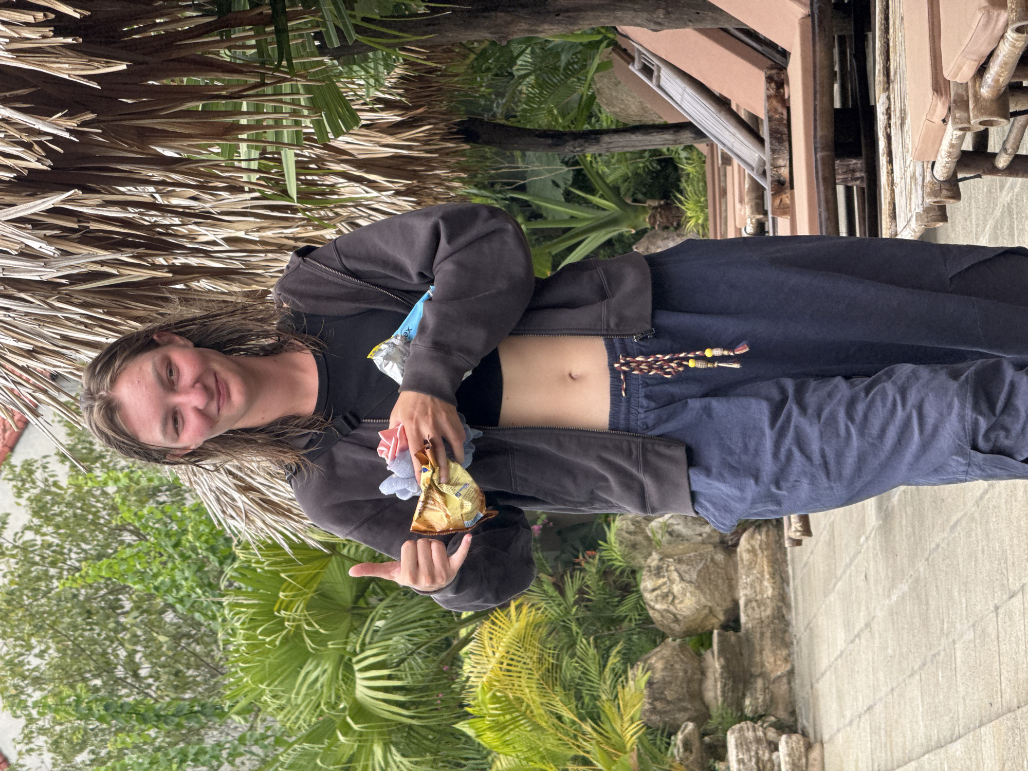 A young person with wet, shoulder-length brown hair is standing outdoors, giving a thumbs-up and holding a small object wrapped in gold foil in their left hand. They are wearing a dark gray zip-up hoodie, a black crop top, and dark blue baggy pants with a braided belt featuring red, yellow, and brown tassels. The person is smiling, looking towards the camera. The background includes lush green foliage, large rocks, and thatched roofing. Several lounge chairs are visible in the middle ground. The ground is paved with light-colored square tiles. The location appears to be a resort or a park-like setting within Nam Hoa Lư Ward, Vietnam.