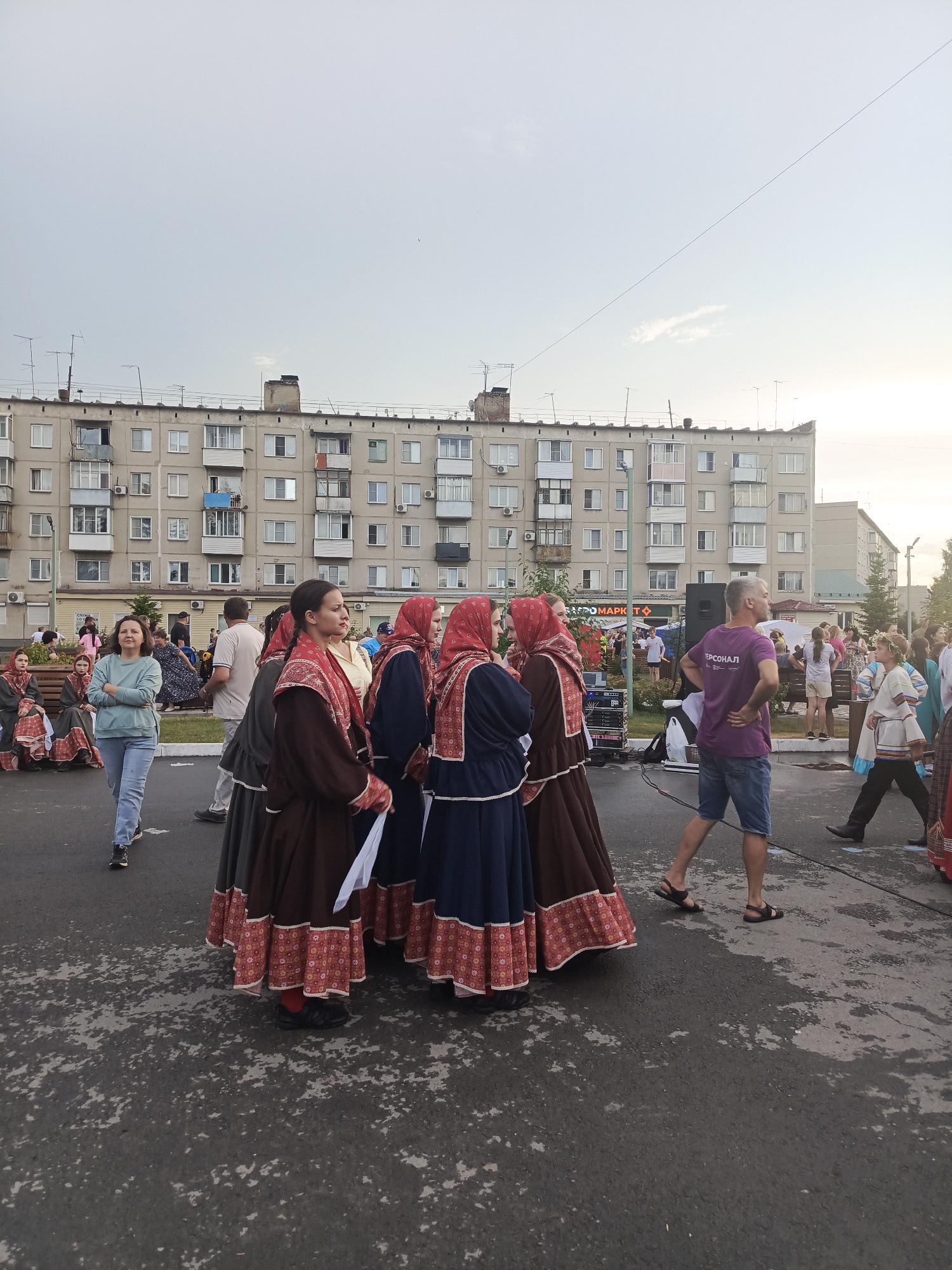 This media file captures an outdoor gathering in a residential area of Krivodanovka, Russia. The scene is set against a backdrop of a typical Soviet-era apartment building, characterized by its monotonous facade and numerous balconies. The sky appears overcast, suggesting it might be late afternoon or early evening, and the weather seems mild.
The primary subjects are a group of people, many of whom are dressed in traditional Russian folk costumes. These costumes feature long, full skirts and ornate embroidery, with women wearing headscarves. The people are standing and walking on an asphalt surface, possibly a street or a public square.
The activity appears to be a cultural event or festival. Some individuals are posing for photographs, while others are engaged in conversation. In the background, there are hints of a stage or performance area with sound equipment, and other attendees are visible, some seated on benches. One notable detail is a sign with Cyrillic text, although it is not entirely legible. A man in a purple t-shirt with Cyrillic text and a woman in jeans and a teal sweatshirt are also prominent figures in the foreground, observing the scene. The overall atmosphere seems relaxed and festive, with a sense of community engagement in a cultural celebration.