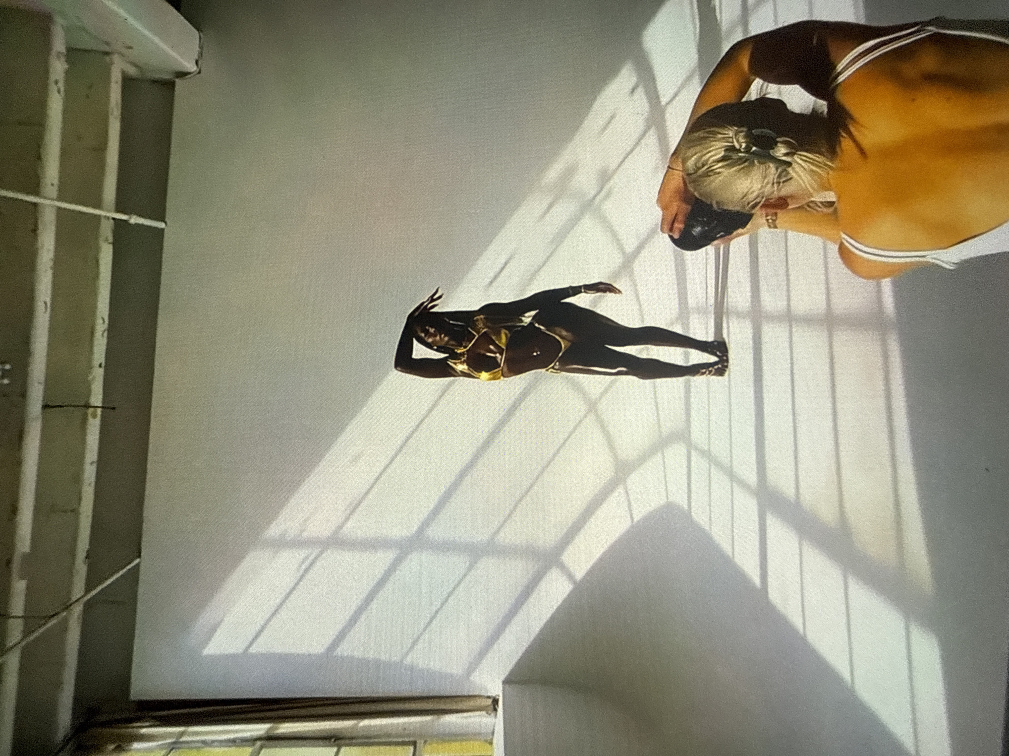 The scene is a photography studio featuring a white seamless wall and floor. A dark-skinned individual, identified as a model, stands centrally, posing in a gold two-piece bikini adorned with body jewelry, including necklaces, a belly button ring, and a waist chain. The model's left arm is raised and bent above their head, while their right arm is slightly extended to the side. Natural light from an unseen window on the left casts sharp, angular, grid-patterned shadows across the white wall and floor. In the lower right foreground, a blonde-haired individual, with their hair styled in a bun, is observed from a rear perspective, actively operating a camera held to their eye, directed towards the model. This individual, presumably the photographer, wears a white tank top. The upper frame of the image reveals an industrial ceiling structure with exposed beams and what appear to be conduits or pipes. There is no visual information within the image to corroborate the specific geographical location as Varna, Bulgaria.