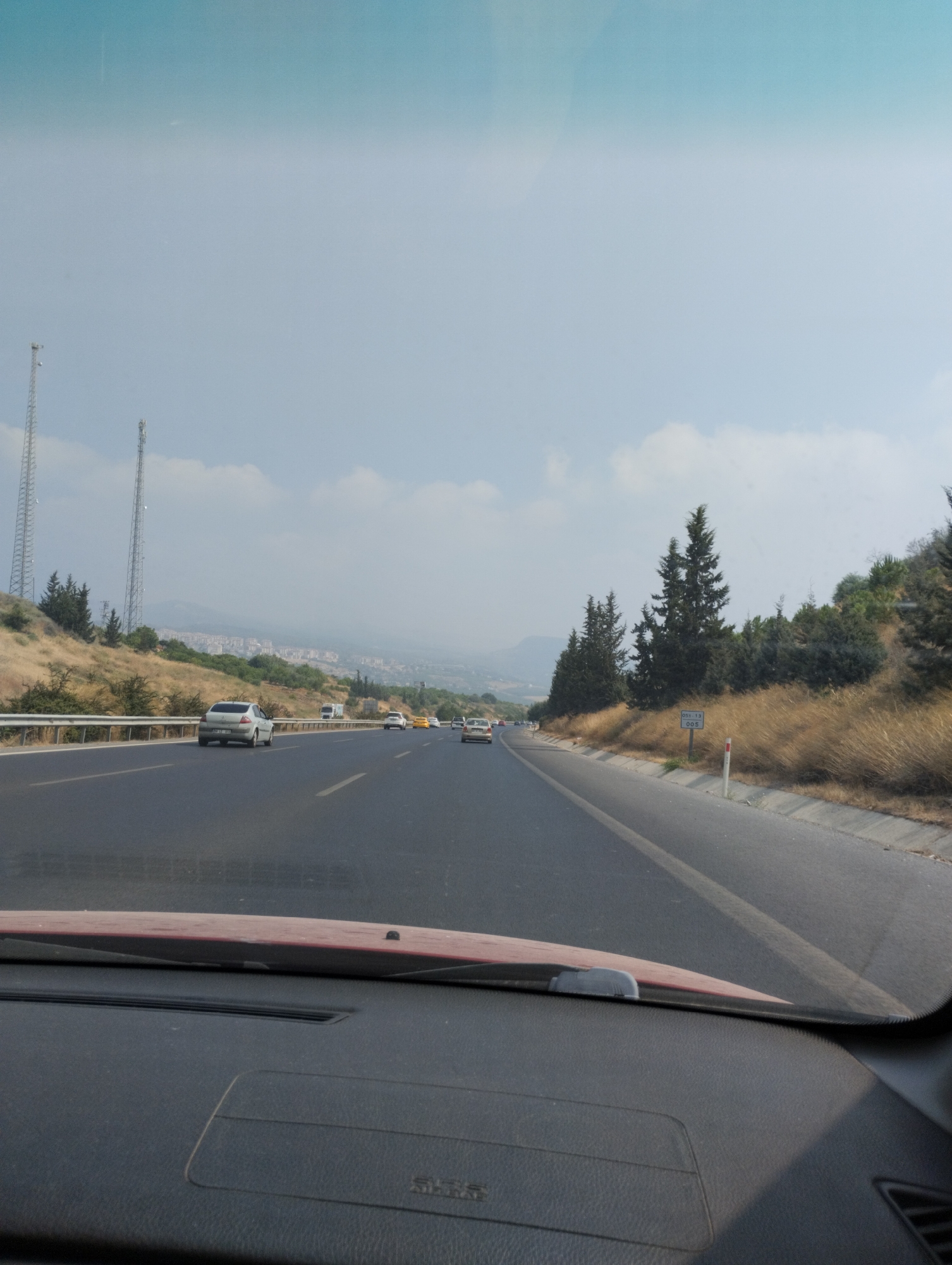 The image shows a view from inside a vehicle driving on a highway in Mezitli, Turkey. The road has multiple lanes and is bordered by dry, grassy hills and scattered evergreen trees. Two tall communication towers are visible on the left side of the road. In the distance, a city or town with buildings is nestled among hills under a hazy sky. Several cars are on the highway, all traveling in the same direction as the viewer's vehicle. The foreground of the image shows the dashboard of the vehicle.