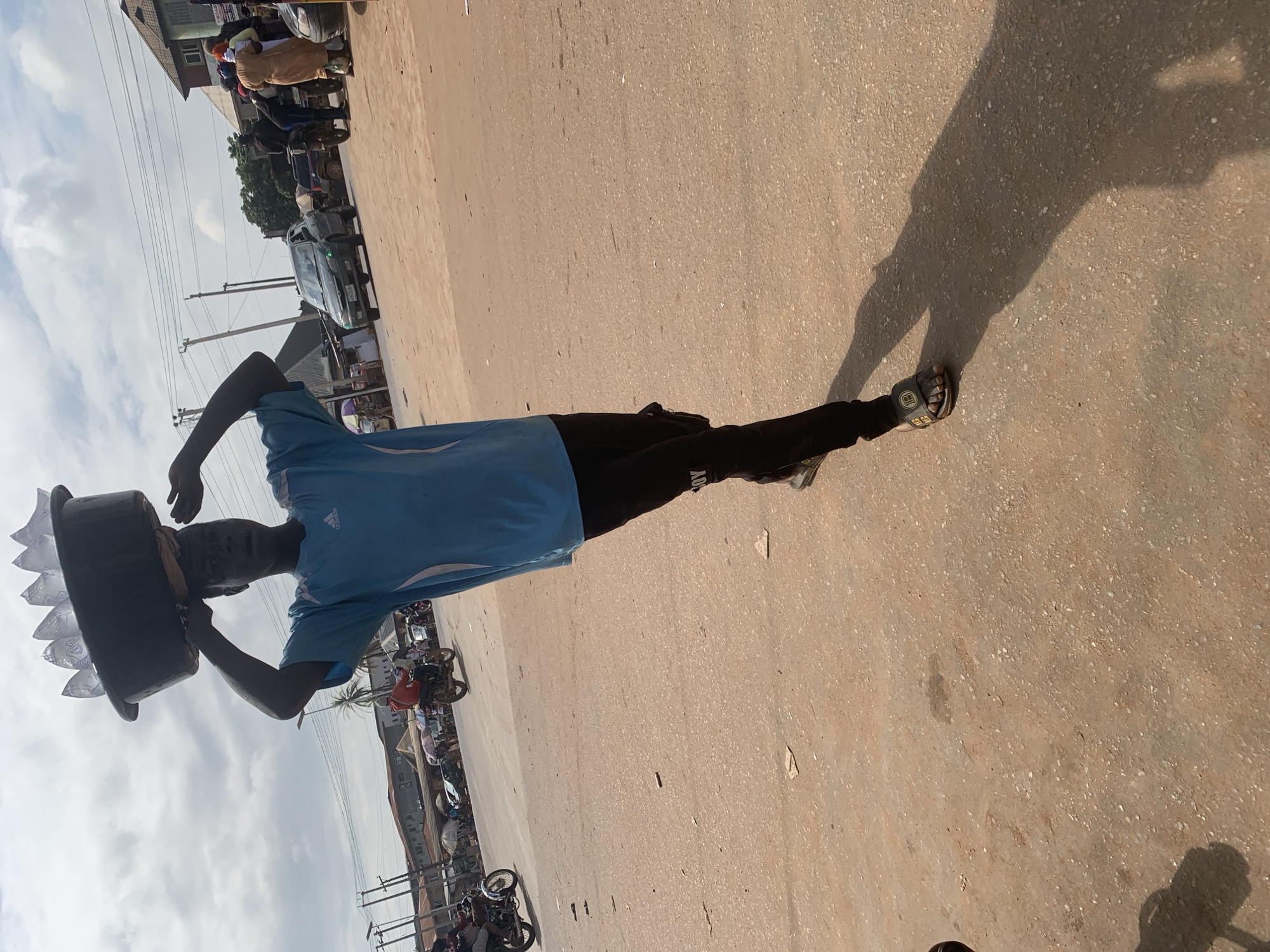 A young man walks down a street in Shagamu, Nigeria, carrying a large basin of water bottles on his head. He is wearing a blue t-shirt and black pants, with sandals on his feet. The street is paved with dirt and gravel, and there are buildings and trees in the background. Several motorcycles are also visible in the scene. The sky is cloudy, and the sun is casting a long shadow of the man on the ground.