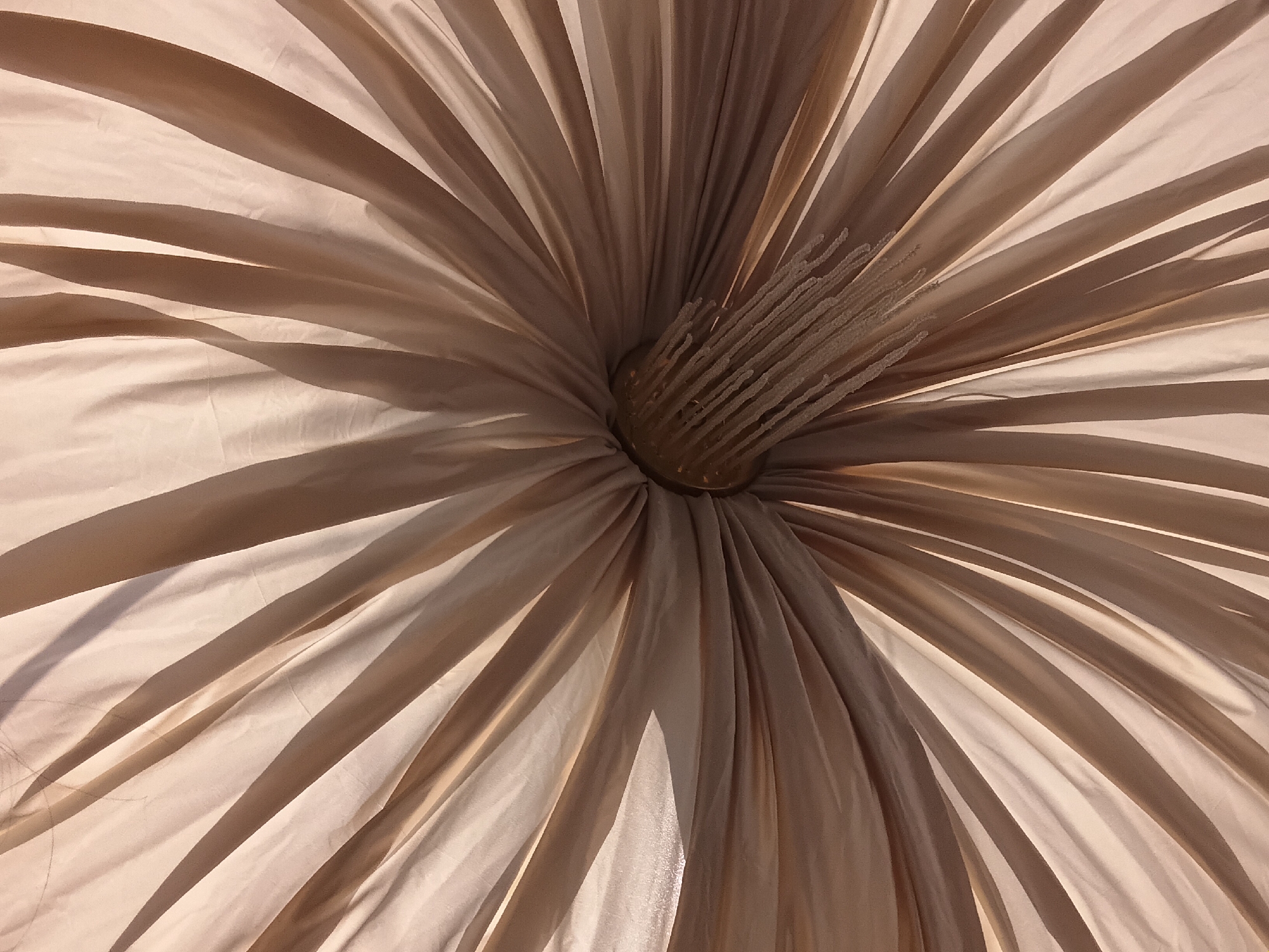 An underside view of a light-colored, fabric umbrella reveals its internal structure. The fabric is pleated and fanned out from a central point, creating a radial pattern of light and shadow. At the center, a metallic mechanism with string-like supports is visible. The lighting suggests an outdoor setting, possibly under sunlight, casting distinct shadows on the fabric. The image does not depict any urban or environmental features specific to the city of Osh, Kyrgyzstan.
