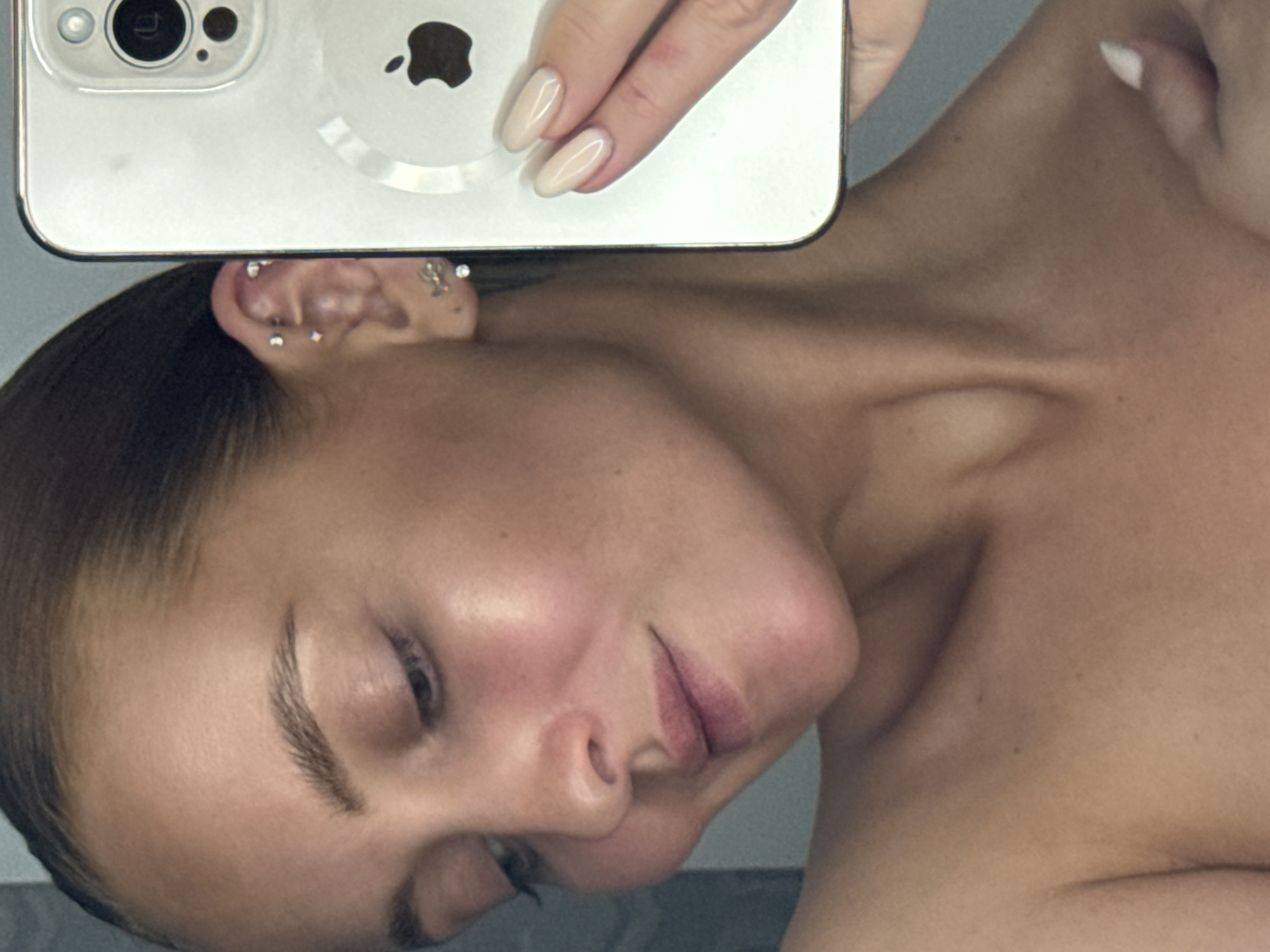 A selfie image shows a person's face and upper torso. The person is holding a silver iPhone above their head and to the side, partially obscuring their ear. The phone's camera and Apple logo are visible. The person has fair skin, arched eyebrows, and a neutral expression. Their hair is slicked back. The image does not provide any information to identify the location as Chaikovsky, Russia, or any other location.