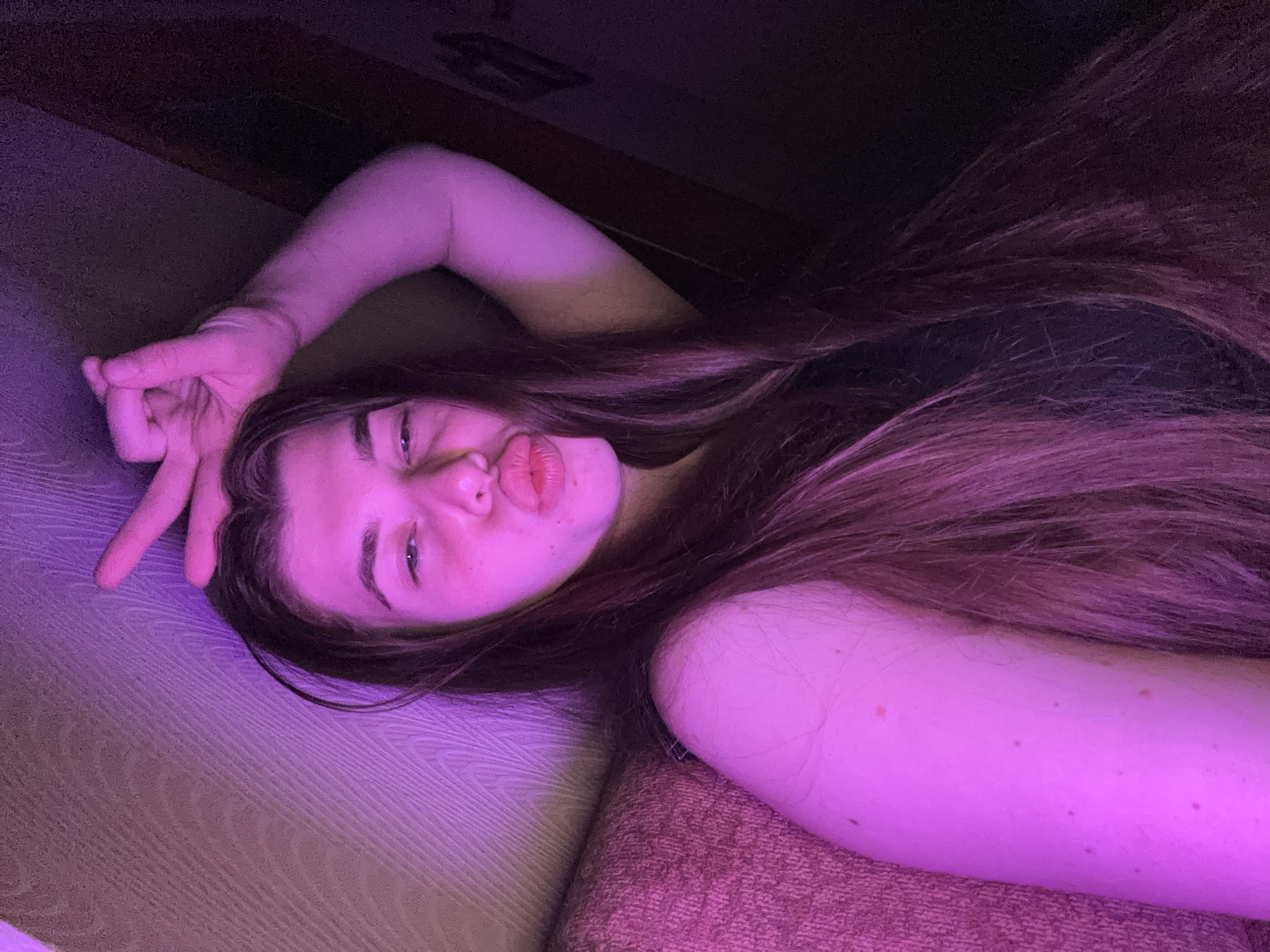 The image shows a young woman lying on her side, looking at the camera. She has long, brown hair and is making a kissy face. Her eyes are open, and she is wearing a black top. She is holding up her right hand in a peace sign. The scene appears to be indoors, with purple lighting. The background is dark, with a hint of what looks like a couch or bed. The woman's skin is illuminated by the purple light, and there are some small marks visible on her arm. The overall mood of the image is playful and casual. The location context is Мещёры, Russia.