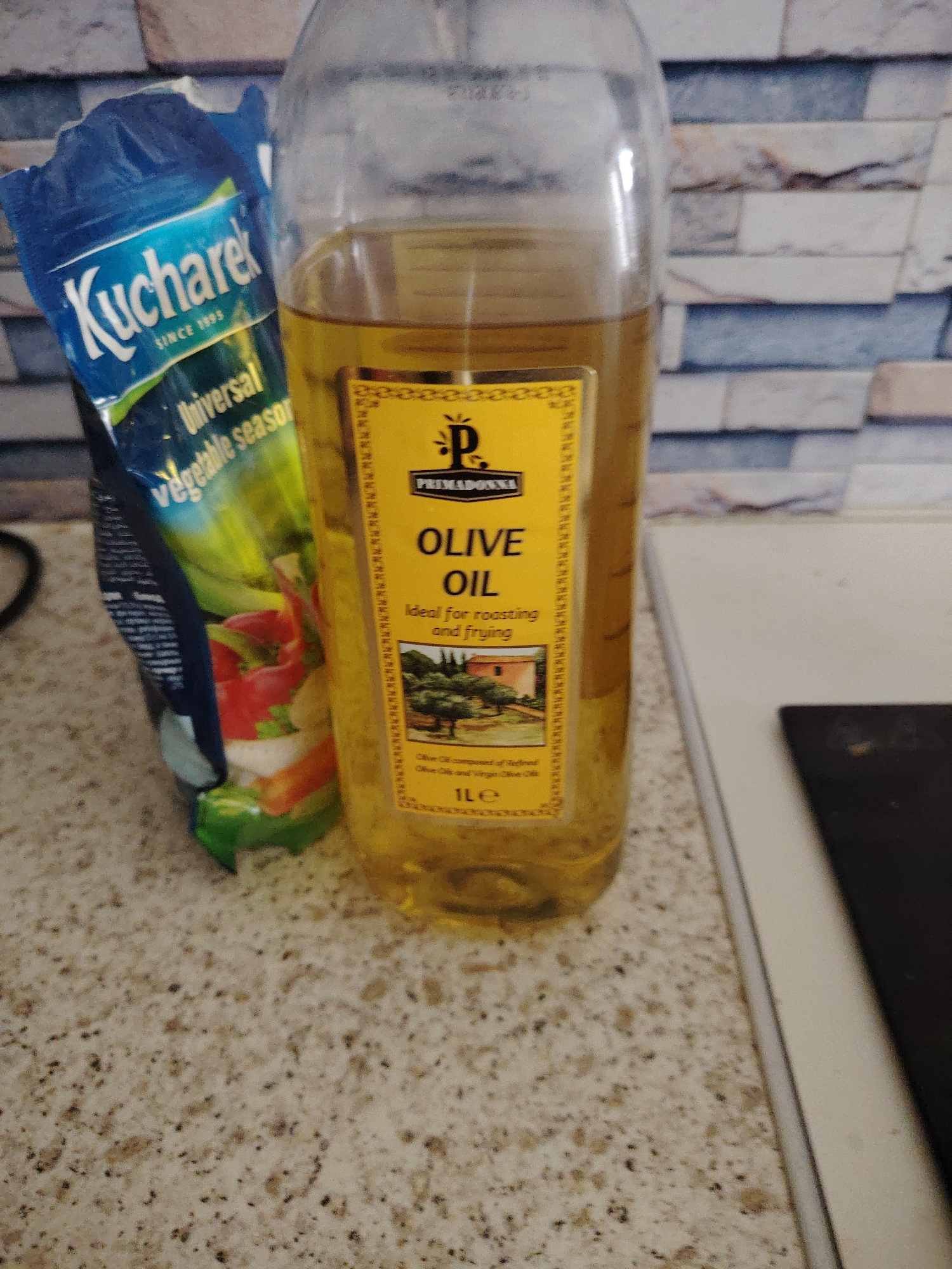 This image, taken in Huntingdonshire, United Kingdom, depicts a kitchen countertop scene featuring two primary items against a tiled wall background. On the left, a partially open blue foil pouch of "Kucharek Universal Vegetable Seasoning" is visible. The pouch features text in white and green, indicating the brand "Kucharek" established "SINCE 1995", and displays an illustration of various vegetables including red peppers, carrots, and white onions. To its right, a clear glass bottle filled with yellow "PRIMADONNA OLIVE OIL" stands upright. The bottle has a yellow label detailing "OLIVE OIL Ideal for roasting and frying", along with a small image of olive trees and a building, and states "1 LE" (1 Liter equivalent). The olive oil is described as "composed of Refined Olive Oils and Virgin Olive Oil". Both items rest on a speckled light-colored countertop. In the background, a wall is tiled with rectangular, irregularly patterned tiles in shades of blue-gray, light brown, and white. To the far right, a segment of a white surface, possibly a stove top or another counter section, is visible, alongside a black, flat object, likely a cooking utensil or burner, separated by a thin metallic strip.