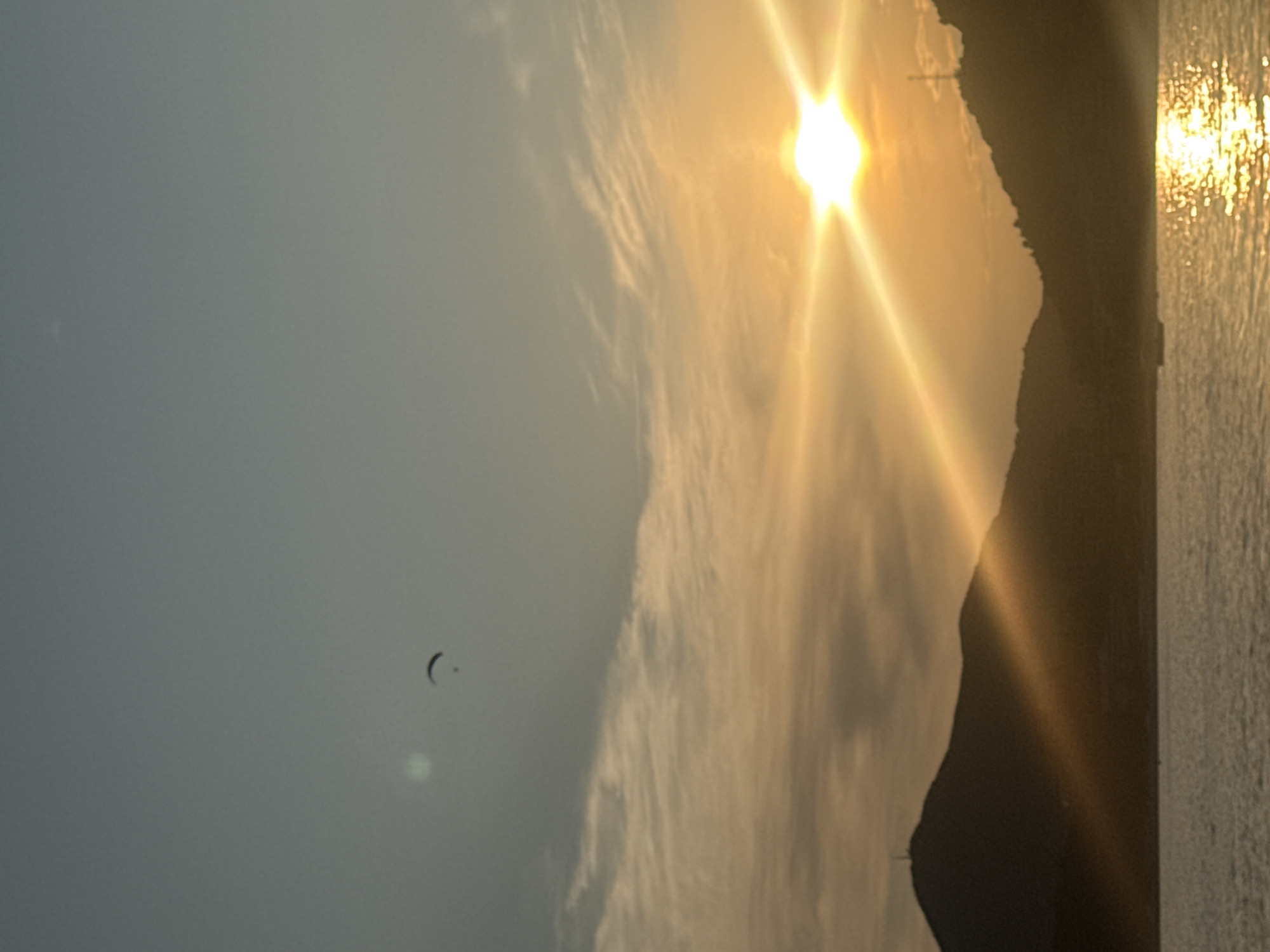 A paraglider is visible in the upper left quadrant of the image, silhouetted against a gradient sky transitioning from clear blue to a warm orange hue. The sun, positioned in the upper right quadrant, emits strong rays that illuminate the sky and cast a golden reflection on the water. In the foreground, dark silhouettes of mountains border a body of water, likely the Adriatic Sea. A structure resembling a tower or antenna is perched atop the leftmost mountain. The water's surface is rippled, with the sunlight creating a bright, shimmering path across it. The overall scene captures a tranquil sunset over a coastal landscape.