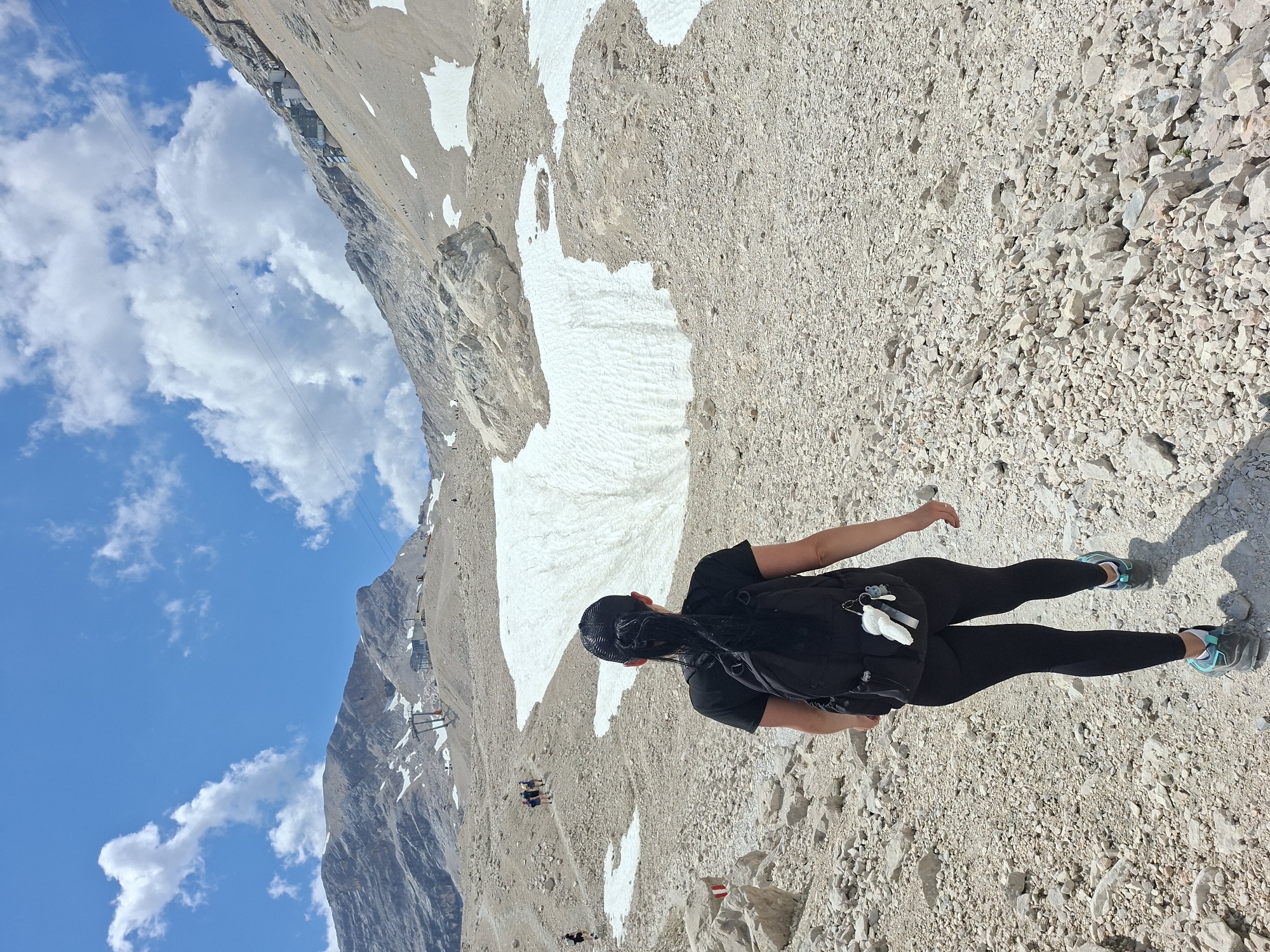The image depicts a person walking on a rocky, mountainous terrain under a bright blue sky with scattered clouds. The person, viewed from behind, is wearing a black t-shirt, black leggings, a black backpack, and hiking shoes. They are walking on a path composed of loose rocks and gravel. In the background, there are more mountains, patches of snow, and what appears to be a ski lift or cable car system. Several other hikers are visible in the distance, ascending the mountain. The environment suggests a high-altitude, summer alpine setting. The location is identified as a mountain area near Grainau, Germany, likely the Zugspitze region given the presence of cable car infrastructure and snow-capped mountains.
