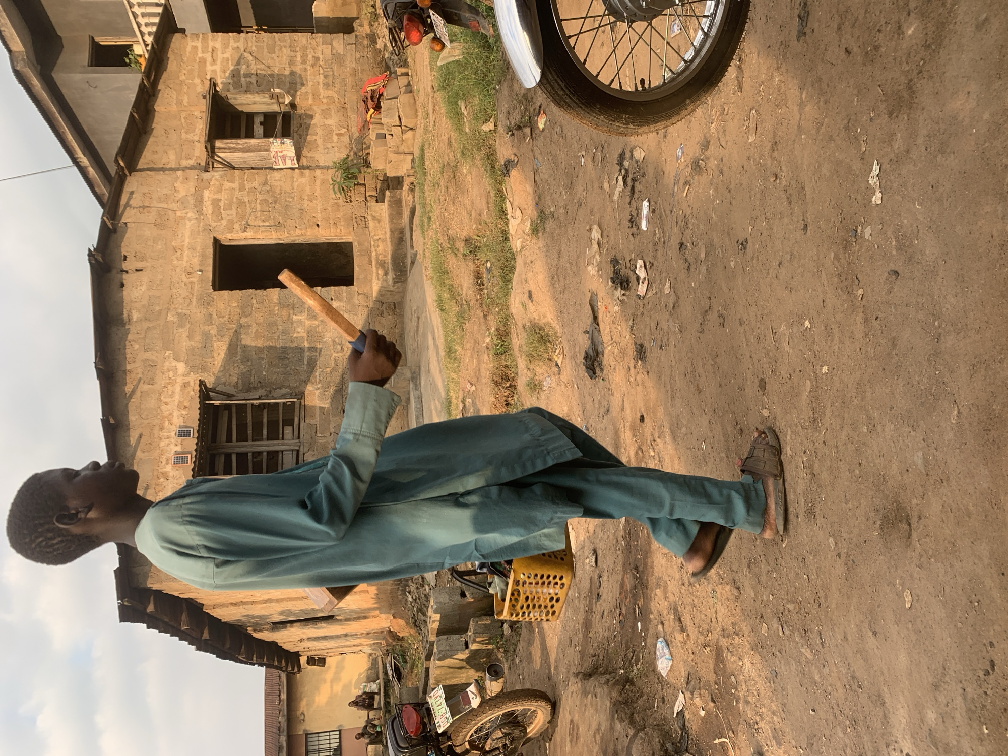 An outdoor, low-angle, eye-level shot shows a young Black male from the shoulders down, walking on a dirt road in Shagamu, Nigeria. He is wearing a teal-colored, loose-fitting robe and pants, and brown sandals. In his right hand, he carries a light brown wooden stick, and on his left hip, a yellow plastic basket is attached. The background features a building constructed from rough-hewn, light brown bricks, with a corrugated metal roof and a simple wooden door. Adjacent to this structure, another building with a lighter, plastered facade is visible. The ground is uneven, covered in dirt, with scattered debris including plastic bags and wrappers. A motorcycle is parked to the left, and another motorcycle's wheel and fender are visible on the right side of the frame. Two other individuals are seated in the background near the brick building. The lighting suggests it is late afternoon with warm sunlight casting long shadows.