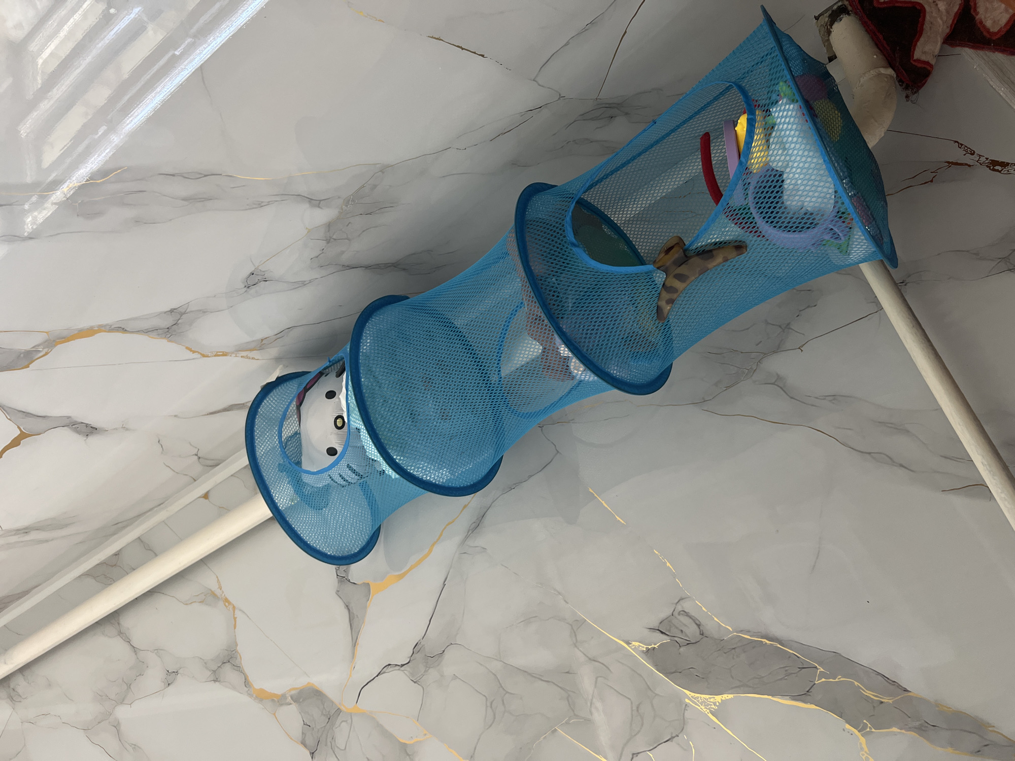 A blue mesh toy organizer hangs vertically against a light-colored marble-patterned wall. Two white poles, likely part of a structure or conduit, are positioned on either side of the organizer and run parallel to each other. The organizer has multiple openings and contains various toys, including a stuffed animal with cat-like features visible in the top opening, a plush toy resembling a giraffe or lizard in the middle opening, and other colorful toys such as rings and what appear to be small balls in the bottom opening. The scene is likely indoors, given the wall finish and the presence of toys. No people are visible in the image. The overall impression is one of organized storage for children's toys in a domestic setting. The location is specified as Бирдик, Kyrgyzstan.