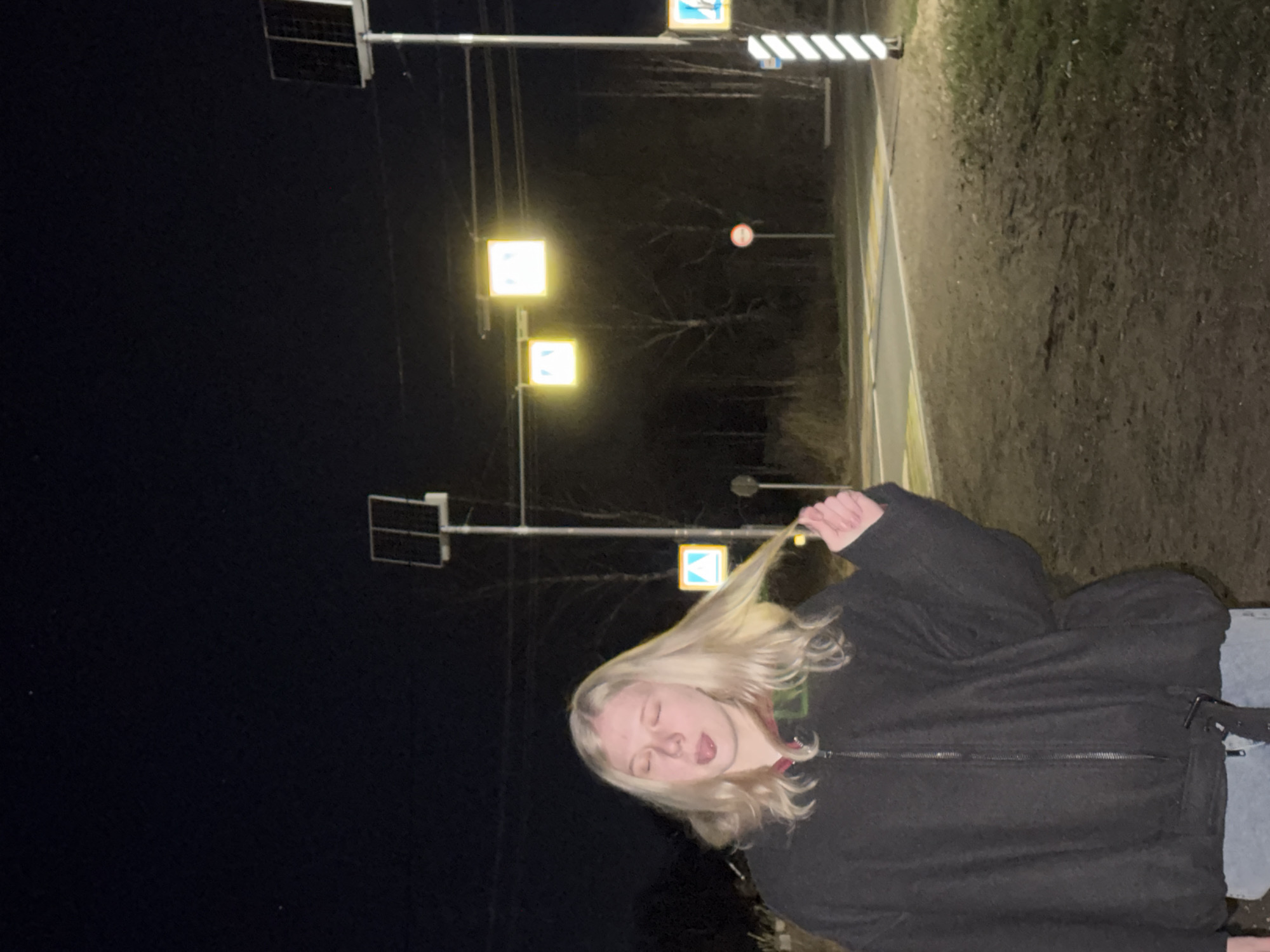 A nighttime scene in an urban environment, likely Unknown, Russia, features a young, fair-skinned person with blonde hair, wearing a dark hooded jacket and jeans. They are positioned near a street, with visible traffic signals and streetlights illuminating the area. The person is facing the camera, holding a strand of their hair, and has their eyes closed and tongue sticking out. The background includes a road, trees, and traffic signs. The overall lighting is low, enhancing the contrast between the illuminated objects and the surrounding darkness.