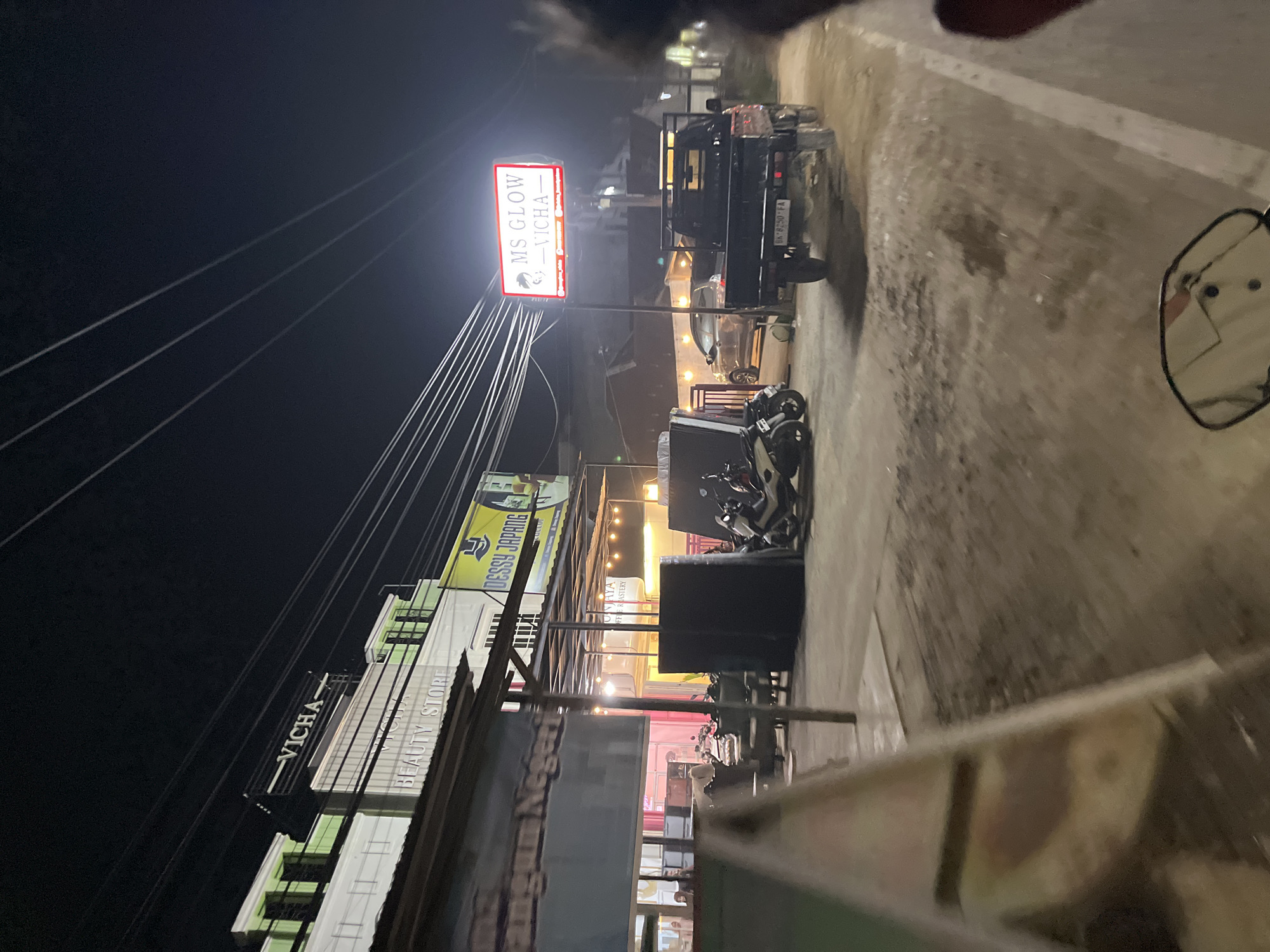 This is a nighttime, outdoor, low-light shot of a street scene in Bundar, Indonesia. The image is rotated 90 degrees clockwise. The foreground is blurry and out of focus, showing a reflective surface that may be a vehicle's mirror, with a faint reflection of a person or people.
The main subject of the image is a street lined with buildings and businesses. There are several signs visible. A prominent, illuminated red and white sign reads "MS GLOW - VICHA". Below this sign, there are many power lines stretching across the frame. Further down the street, a building has a black sign with white lettering that says "VICHA". To the right of that, another building has a sign that reads "BEAUTY STORE". There is also a yellow sign with dark text that appears to be an advertisement, possibly for "DESSY JEPANG".
Several vehicles are parked along the street. A black pickup truck is parked near the "MS GLOW" sign. Further down, there are a few motorcycles. The buildings appear to be commercial establishments, with lights on inside some of them, suggesting they are open. The overall atmosphere is that of a lively, albeit dimly lit, commercial area at night. The road surface appears to be paved but shows some wear and tear. The weather is unclear, but the lighting suggests it is nighttime. There are no discernible emotions conveyed in the image due to the lack of clear human subjects and the low-light conditions.