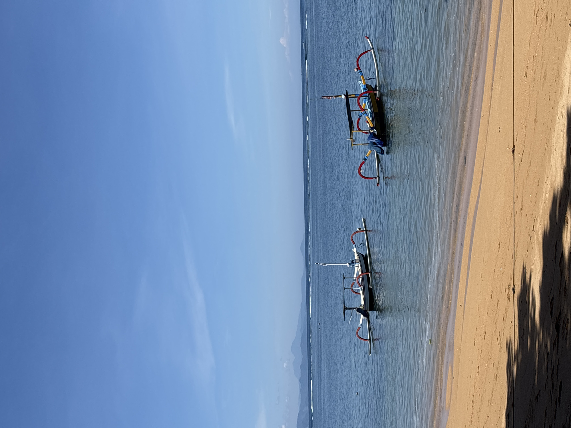 The image shows a coastal scene in Denpasar, Indonesia. Two traditional outrigger boats are anchored near a sandy beach. The water is clear and blue. The sky is clear with some clouds. There is no one on the beach or boats. The boats have distinctive curved outriggers, painted red and silver. A mountain range is barely visible in the background.
