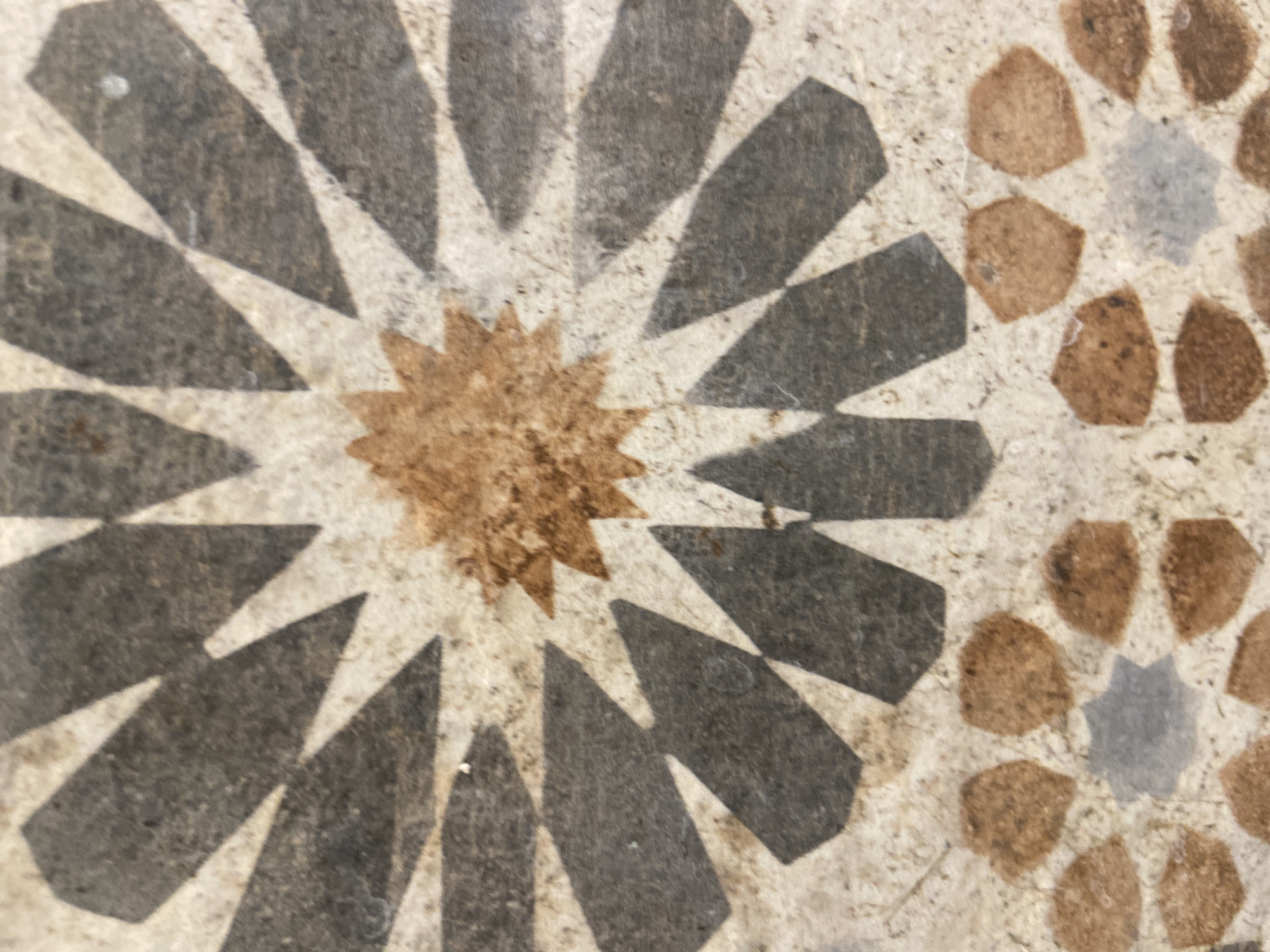 The image displays a close-up view of a decorative tile with a geometric pattern. The pattern features a central star-like motif with brown and dark gray elements radiating outwards. Below this central design, a smaller circular arrangement of brown shapes surrounds a central eight-pointed gray star. The tile appears to be made of stone or ceramic with a slightly textured, matte surface, showing some speckling and minor imperfections. The lighting is diffused, casting soft shadows that highlight the relief of the pattern. The context of "Unknown, Unknown" indicates that the specific location of this tile is not provided.