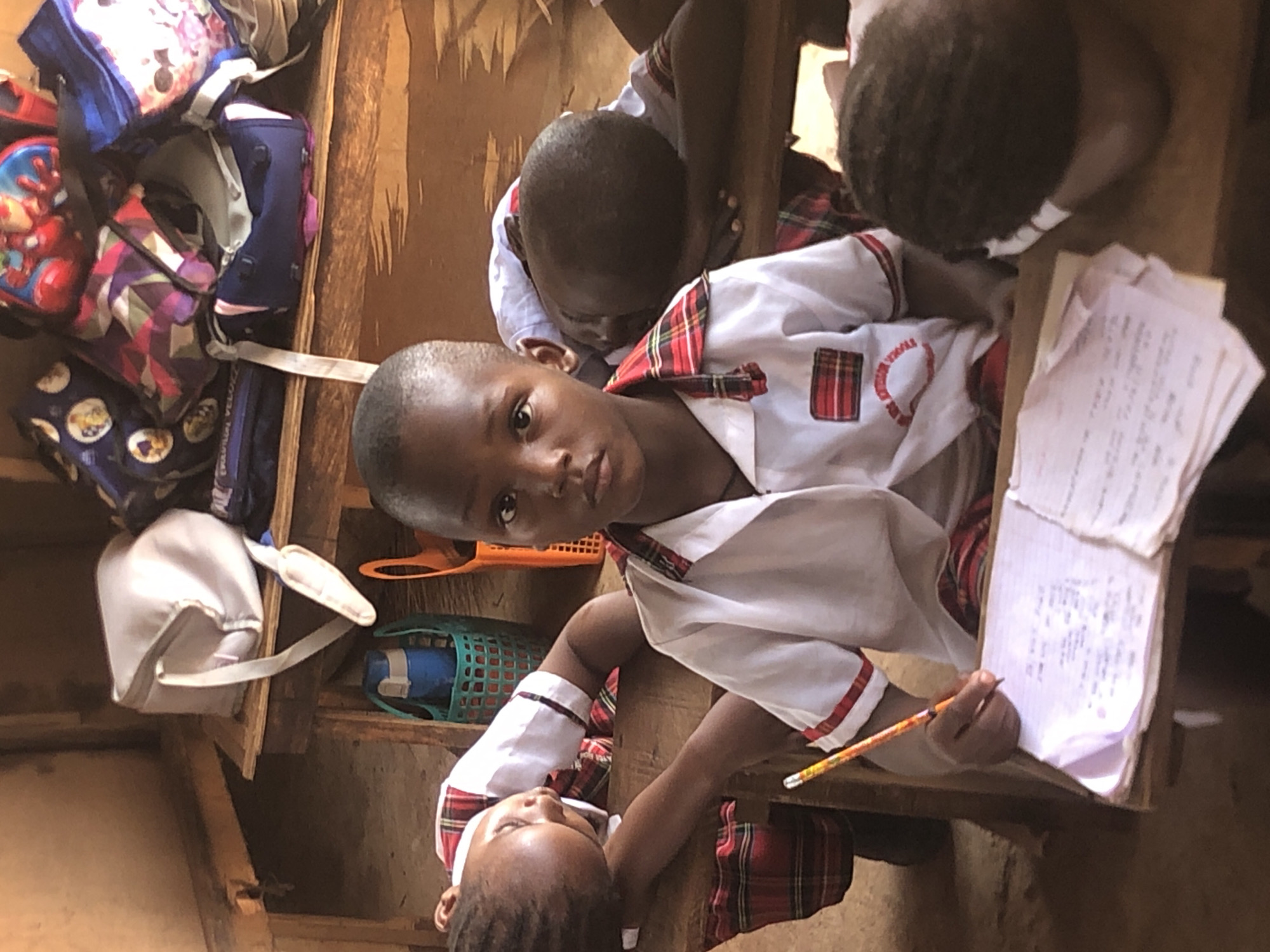 The scene depicts four young children in a classroom setting in Ukwu-Abwa, Nigeria. All the children are wearing white shirts with red and green plaid accents, indicating school uniforms. One child, centered in the foreground, is looking directly at the camera while holding a pencil and writing on a piece of paper. Two other children are partially visible behind the child in the foreground, and the fourth child is resting on the desk. There are bookbags in a storage area, and personal items on shelves beside the desk. The classroom appears to be modestly furnished.