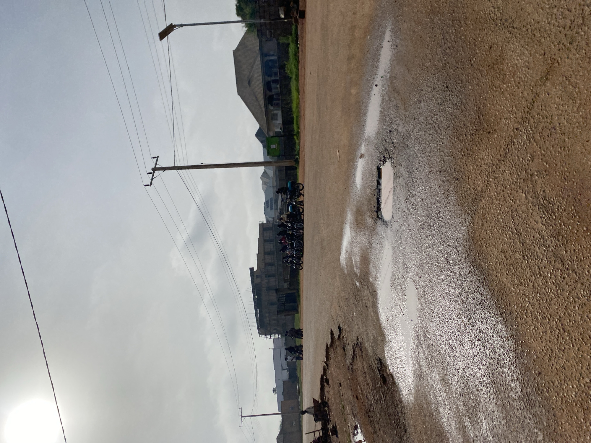 The image depicts a street scene in Kwang, Nigeria, likely in the late afternoon given the overcast sky and diffused sunlight. The perspective is from ground level, looking towards a row of buildings and a street.
The primary subjects are the road, buildings, and several motorcycles. The road surface is asphalt, characterized by a rough texture and the presence of several puddles, suggesting recent rain or damp conditions. One large puddle with a distinct rectangular shape is particularly prominent, reflecting the sky. There are also several potholes and rough patches on the road.
In the background, a line of buildings of varying heights and architectural styles can be seen. Some appear to be residential or commercial structures, with multiple stories and balconies. One building is under construction. Amidst the buildings, green vegetation is visible, indicating a potentially tropical or sub-tropical environment.
Several motorcycles are parked or being ridden along the road, forming a small procession. People are visible on and around the motorcycles, though their features are not clearly discernible due to the distance and resolution. They appear to be dressed in everyday clothing.
Overhead, power lines crisscross the sky, supported by utility poles. A lamppost is also visible. The sky is cloudy and a bright, hazy sun is partially visible in the lower left corner. The overall impression is one of an active, everyday street scene in a Nigerian town. There is no visible text in the image.