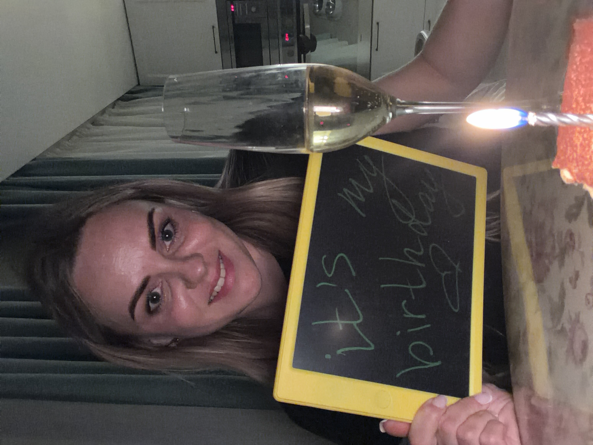 A woman is holding a birthday cake with a lit candle and a glass of champagne. She is also holding a chalkboard that says "It's my birthday". The photo was taken indoors with a kitchen visible in the background, implying it may have been taken in the city of Votkinsk, Russia.
