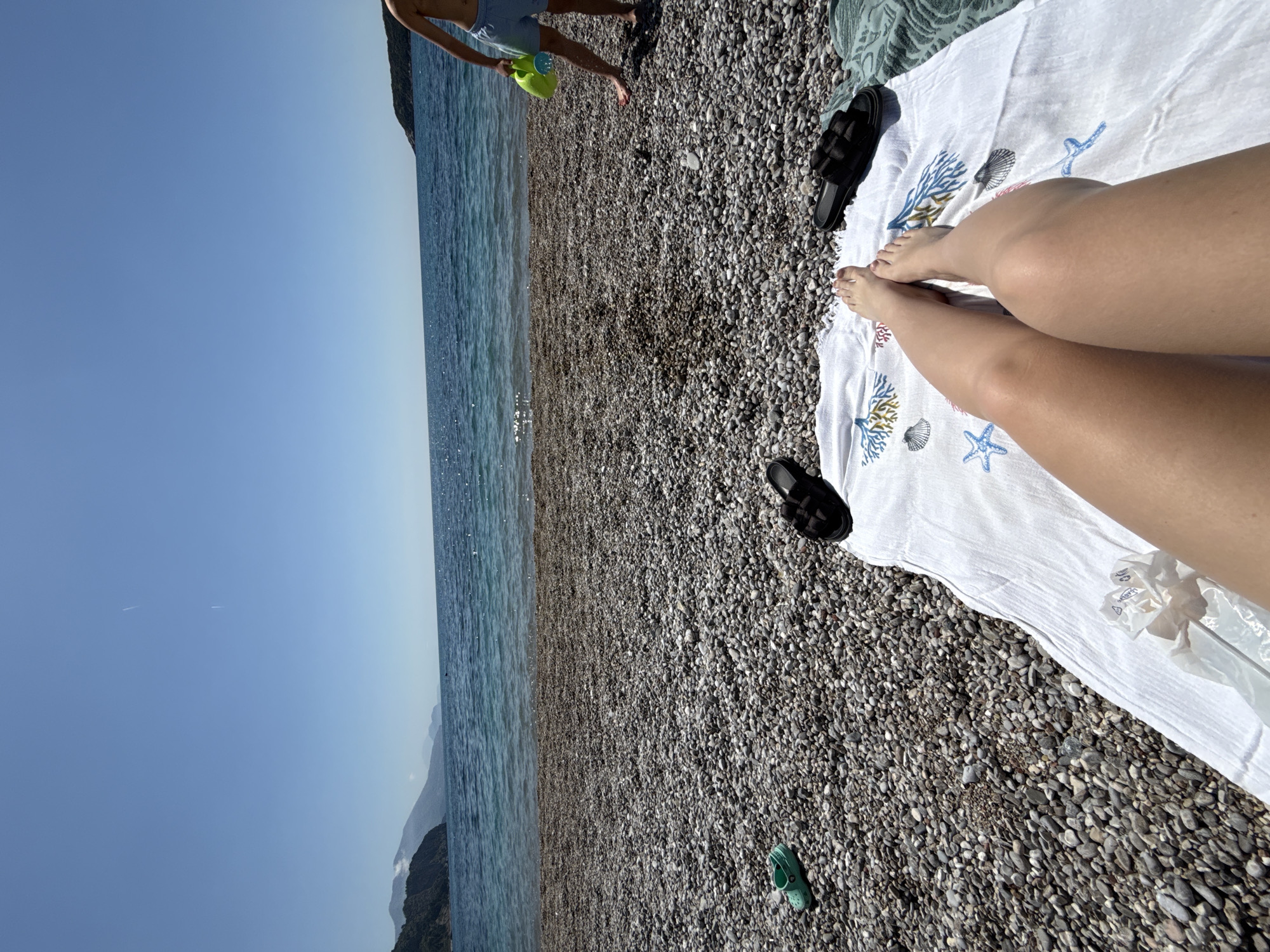 The image captures a scene at a beach in Prijevor, Montenegro. The environment consists of a pebble beach meeting the clear turquoise sea under a bright blue sky.
A person is lying on a white beach towel with sea-themed patterns. The person's bare legs are visible on the towel, indicating relaxation. A pair of black sandals sits near the person's feet, suggesting leisure.
In the background, another individual, presumably male, is seen standing near the water's edge, wearing blue shorts and holding a green watering can. A green Croc shoe lies on the pebble beach. The presence of these objects and individuals points to a casual beach outing.