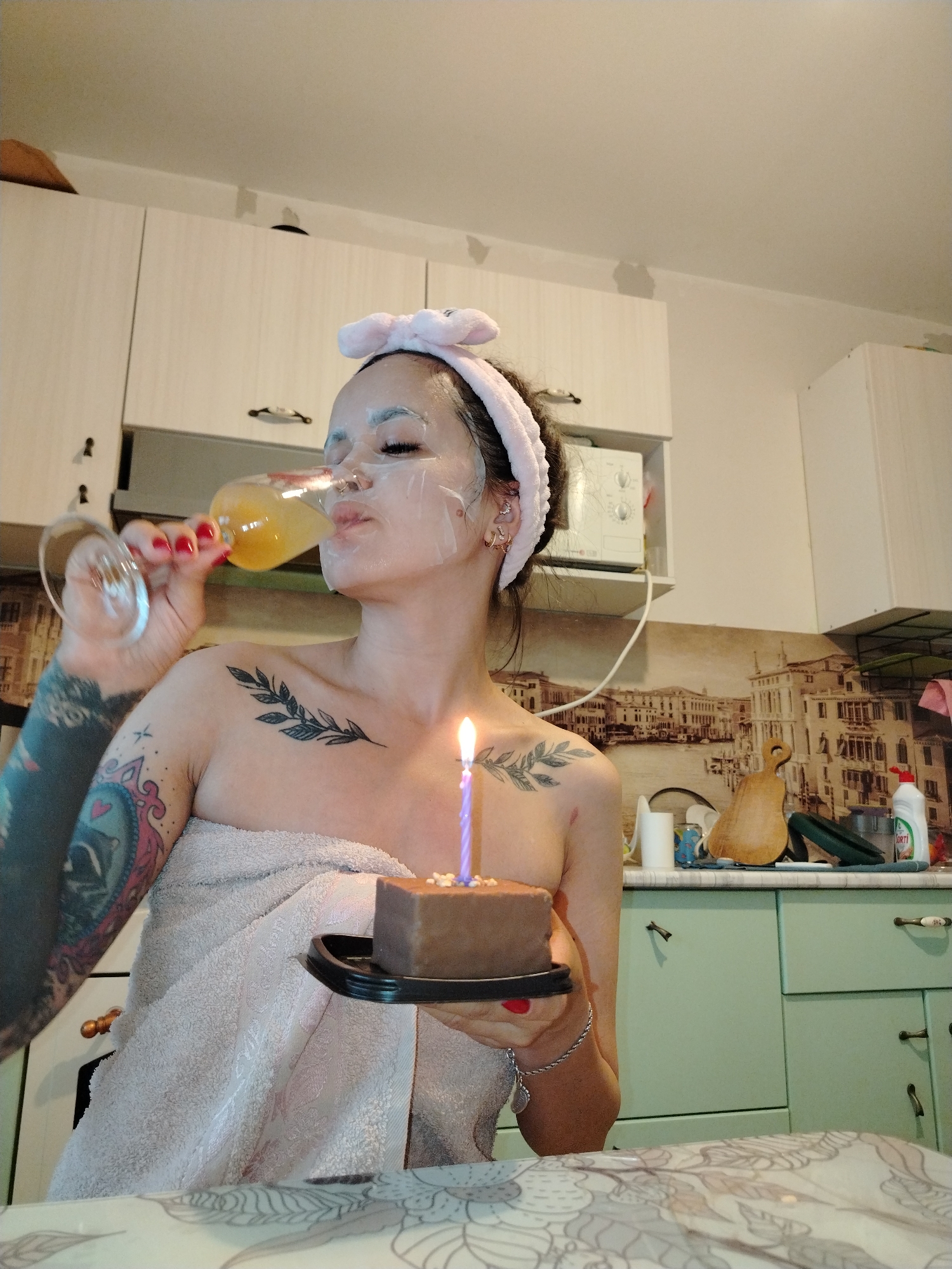 A woman, wearing a face mask, a pink headband, and wrapped in a towel, is holding a glass of amber liquid and drinking from it. She is also holding a small chocolate cake with a lit candle. Tattoos are visible on her left arm and shoulder. She is in a kitchen with light-colored cabinets and a pastel green countertop. There is a backdrop with a painting of a European city. The image appears to be taken indoors, possibly in a residential setting. There is no information in the image to determine the specific location in Noviy, Russia.