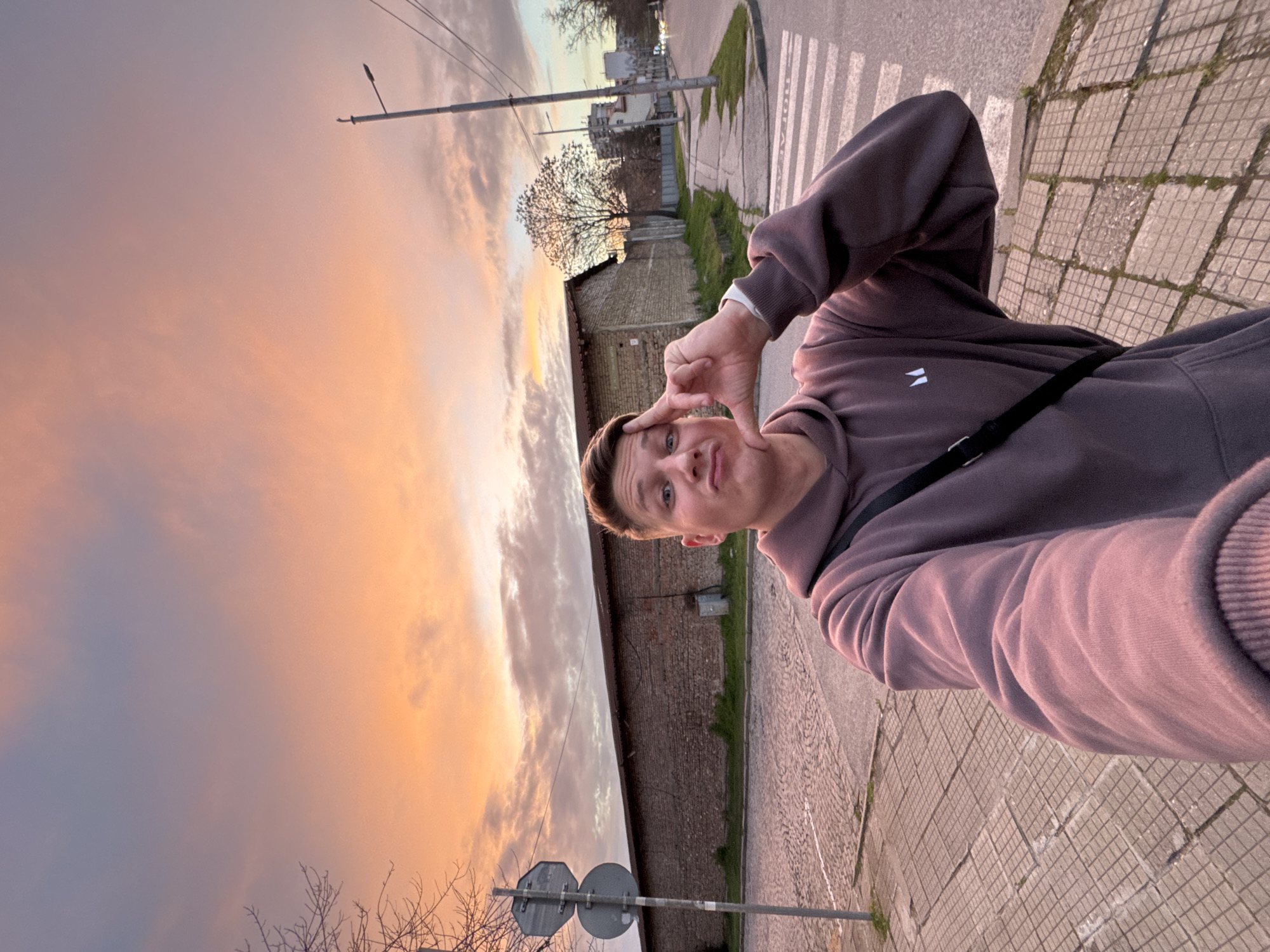 A young Caucasian male is standing on a tiled sidewalk next to a street in Plovdiv, Bulgaria, posing for a selfie-style photo. He is wearing a mauve hoodie and has a black strap across his body. His right hand is raised, forming a square shape with his fingers near his face, which is slightly tilted.
Behind him, a long brick building runs parallel to the street, with some greenery visible between the sidewalk and the building. Further in the background are trees and some buildings suggesting an urban area. In the sky, there's a vibrant sunset with shades of orange and gray. A street lamp is seen on the left side of the photo.
At the bottom left corner, street signs are mounted on a pole, and on the right side of the street, a pedestrian crosswalk is visible.