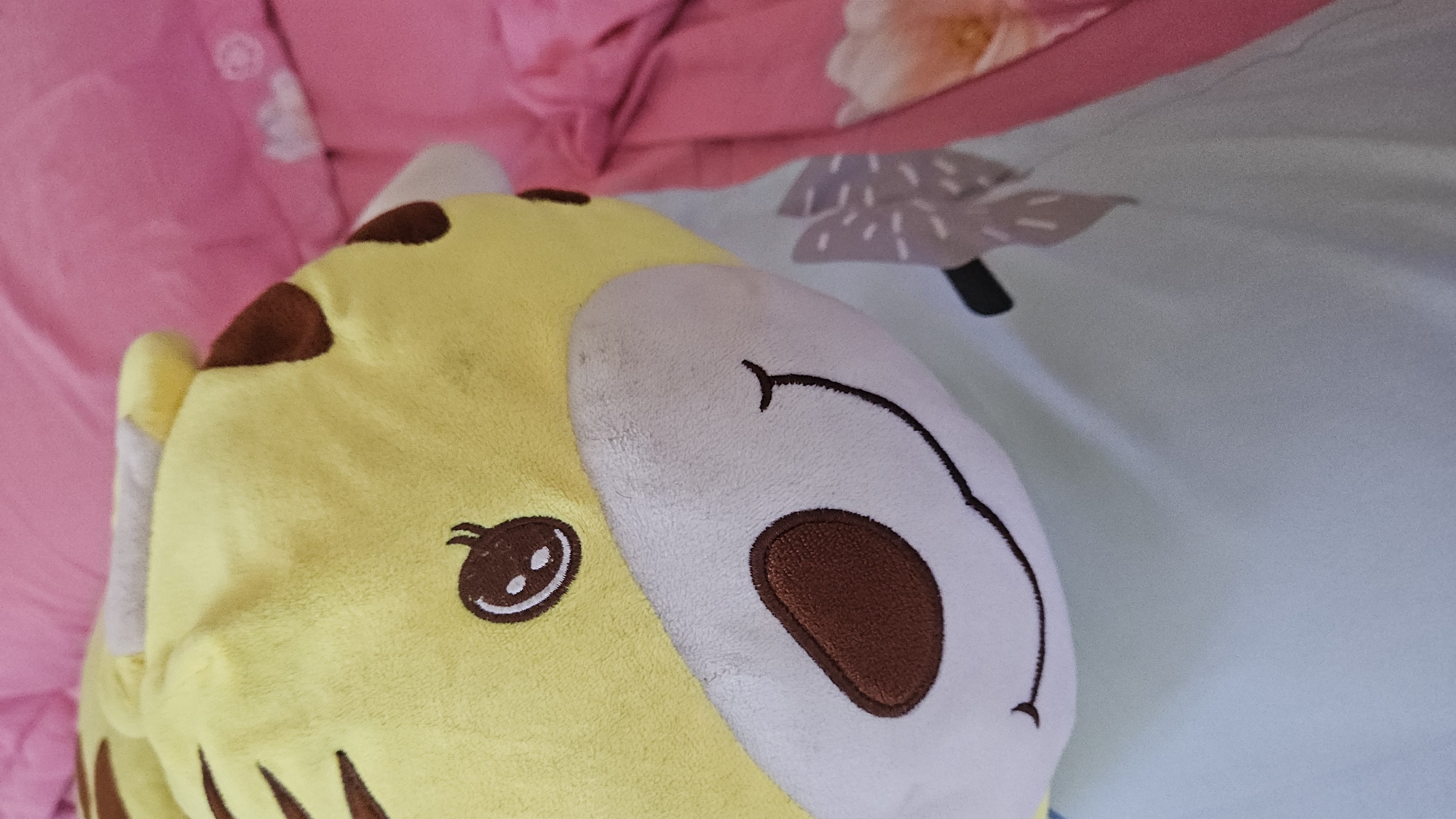 A stuffed animal with a yellow and white face lies on bedding. The left side of the face is yellow with brown spots, an embroidered eye, and brown stripes. The right side is white with a brown embroidered nose and a stitched-on smile. The bedding has a pink sheet on the left side and a blue sheet with a tree design on the right. The location is unknown.