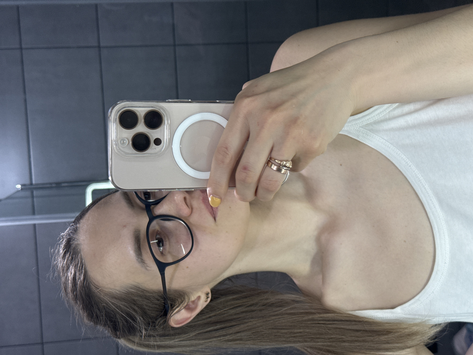 A woman is taking a selfie in front of a bathroom mirror with dark gray tiles. She is holding a smartphone with a clear case, three camera lenses, and a white magnetic ring on the back, partially obscuring her face. The woman is wearing black framed glasses and a white tank top. She has shoulder-length brown hair. She is wearing rings on one of her fingers. The location is likely an indoor bathroom, potentially in a private residence. The city of Chaikovsky, Russia is not verifiable based on the image.