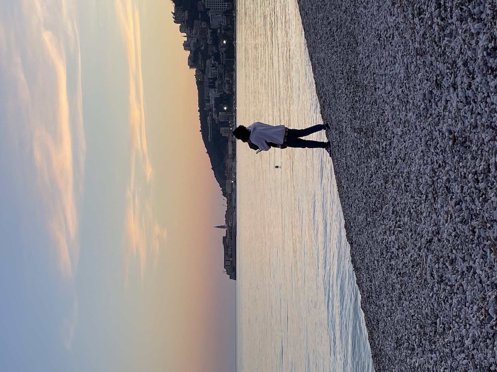 The image depicts a coastal scene at dusk in Budva, Montenegro, observed from a pebbled beach. The sky graduates from pale blue at the zenith to a soft orange and pink hue at the horizon, featuring sparse, elongated clouds. The calm, reflective sea surface shows subtle ripples, mirroring the sky's colors. In the mid-ground to the left, the Old Town of Budva is visible on a headland, characterized by its historical buildings and a prominent bell tower identified as the Church of St. John. To the right of the Old Town, modern buildings are built into a dark, tree-covered hillside, with distant lights beginning to illuminate.
In the foreground, a single individual stands on the grey-white pebbled beach, facing away from the viewer and towards the sea and Old Town. The individual is dressed in a light-colored hooded sweatshirt, dark trousers, and dark athletic shoes with colored accents. They are holding a small, dark-haired child, also facing away, in their arms. The individual's left hand holds a dark-colored mobile phone, raised towards the horizon. A dark, possibly spherical buoy floats in the water approximately midway between the individual and the Old Town.