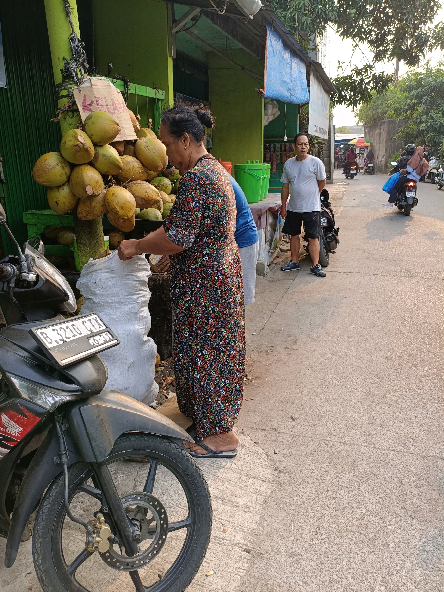 In Tangerang, Indonesia, on a sunny day, a woman in a colorful floral dress is seen bagging coconuts from a large pile. She stands next to a green structure adorned with many coconuts, some still attached to branches. A white sack lies open on the ground, ready to be filled. Beside the sack, a black motorcycle with a license plate reading "B 3216 CTX" is parked. In the background, a man in a gray t-shirt and black shorts is visible, along with other people and motorcycles on a street lined with trees and buildings. The scene depicts a typical street-side market activity in Indonesia.