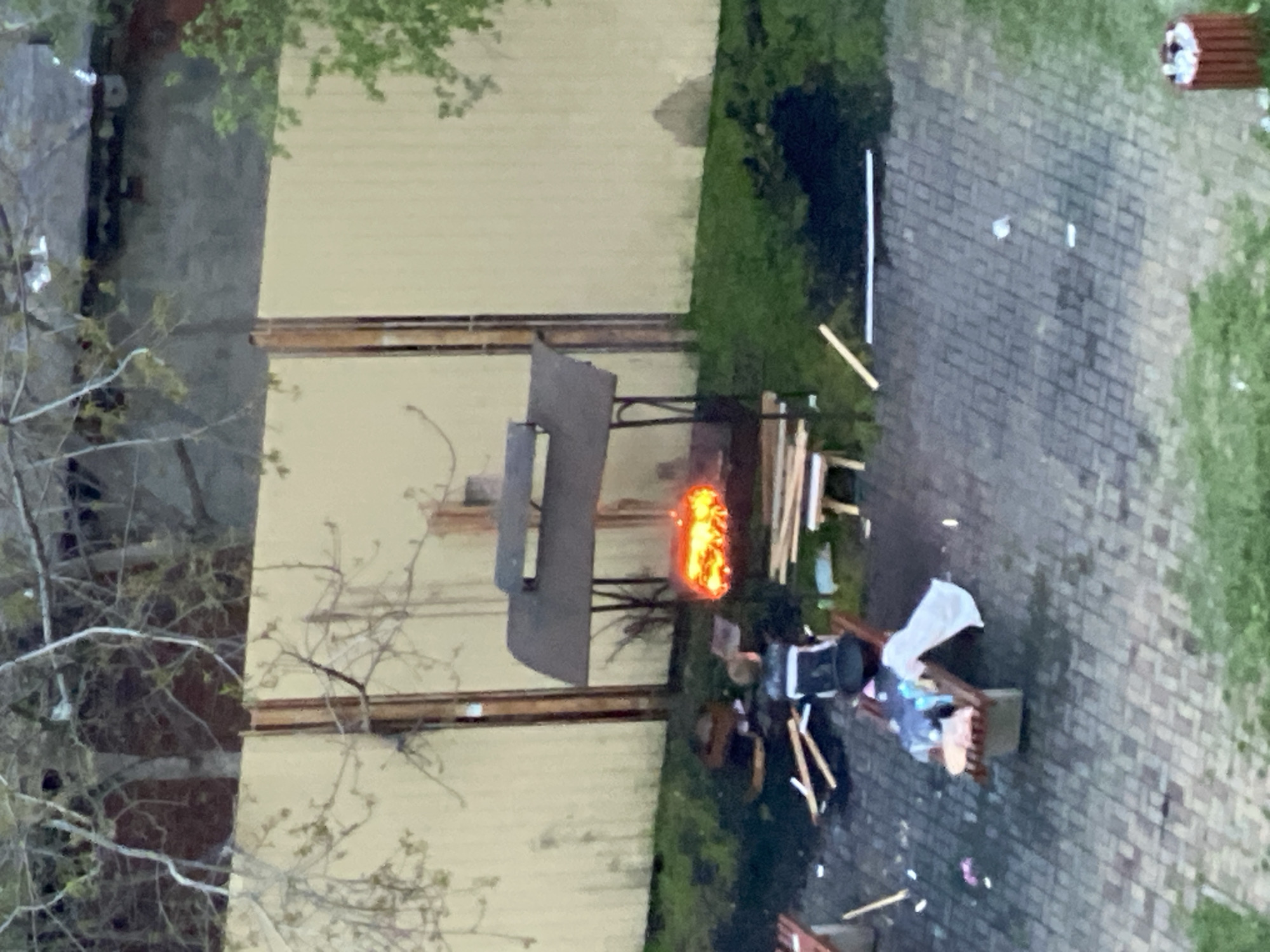 The image appears to be taken from an elevated perspective, possibly from a window. The scene shows an outdoor area adjacent to a light-colored building, with a brick-paved ground extending into a grassy area. In the center of the frame, there is a structure that seems to be some kind of grill or cooking setup with a fire burning inside. Adjacent to the structure, there is debris including wood, a garbage can, and other objects scattered around, suggesting some form of active burning or recent cleanup. There is a long section of the same structure next to it, possibly additional metal pieces of the first one. The environment is a mix of urban or suburban, with visible buildings and paving.