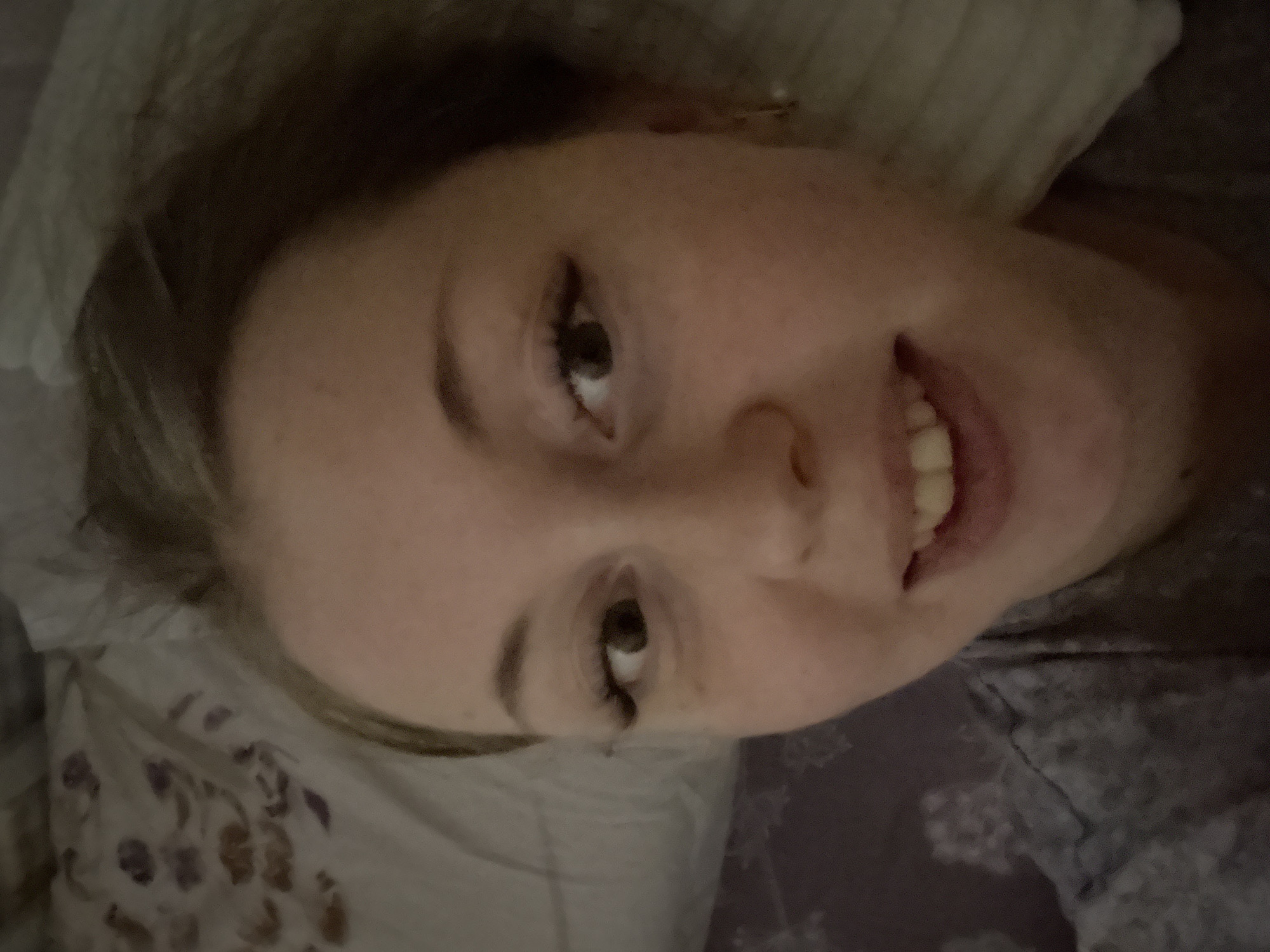 A close-up, tilted portrait captures a person with fair skin, light brown hair, and visible teeth, positioned on what appears to be a bed. The person is smiling, with eyes looking upwards and to the left. The background includes a floral patterned pillow and a dark, patterned bedspread. The location is unknown.