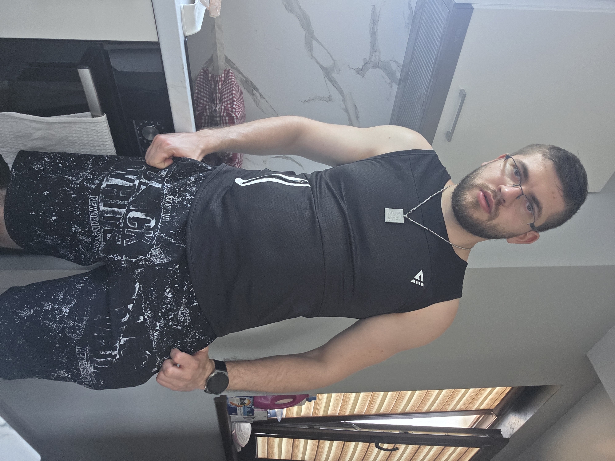 A man is standing in what appears to be a kitchen. He is wearing glasses, a black sleeveless shirt, and black shorts with white lettering. He has a beard and a necklace with a pendant. He is gripping the sides of his shorts with both hands. The kitchen has white cabinets, a microwave, a towel, and a window with blinds. The exact location in the city of Yagodovo, Bulgaria cannot be determined from the image.