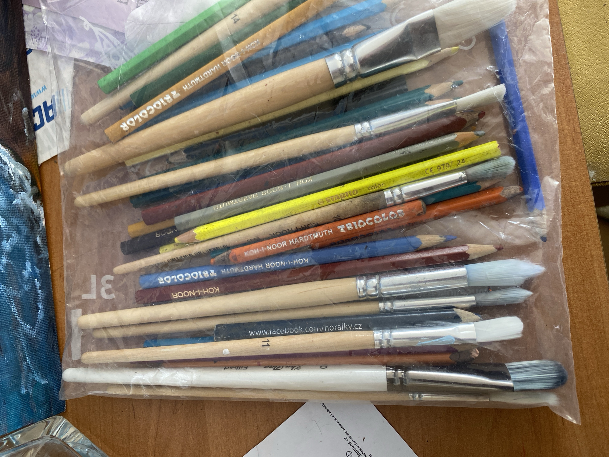 The image shows an assortment of art supplies, including paintbrushes and colored pencils, contained within a clear plastic bag. The items are arranged loosely within the bag, with some brushes standing upright and others lying horizontally. The paintbrushes have wooden handles of varying colors and lengths, and their bristles appear to be made of natural hair or synthetic fibers. Some of the bristles are white, while others are gray or black. The colored pencils are also of various colors and brands, with some featuring text such as "Koh-i-Noor" and "Triocolor" visible on their barrels. The plastic bag itself is slightly crinkled, suggesting it has been handled. The background consists of a wooden surface, likely a table, with a partially visible painting in the upper left corner. There are no people or animals present in the image. The scene appears to be an indoor setting, likely a home studio or art supply store. The city of Bílina, Czechia, is not explicitly depicted or indicated in the image.