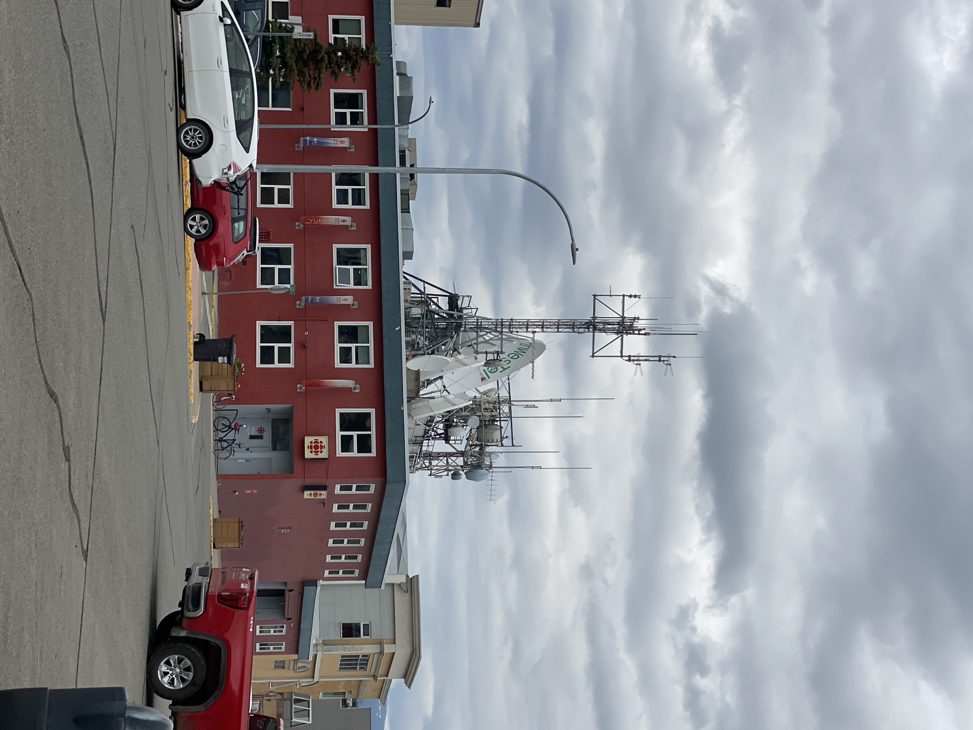 The image depicts a street scene in Whitehorse, Canada, characterized by a red building adorned with a Canadian Broadcasting Corporation (CBC) logo and a complex array of communication antennas and satellite dishes on its roof. The building is situated in an urban setting, with other buildings visible in the background, including a tan and grey structure to the right. In the foreground, a grey asphalt parking lot features several parked vehicles: a white hybrid car, a red station wagon, and a red pickup truck. Two bicycles are parked near the entrance of the red building. The sky is overcast with dark, heavy clouds, suggesting an impending or recent rainfall. The scene is captured from a street-level perspective, looking towards the building and its rooftop equipment.