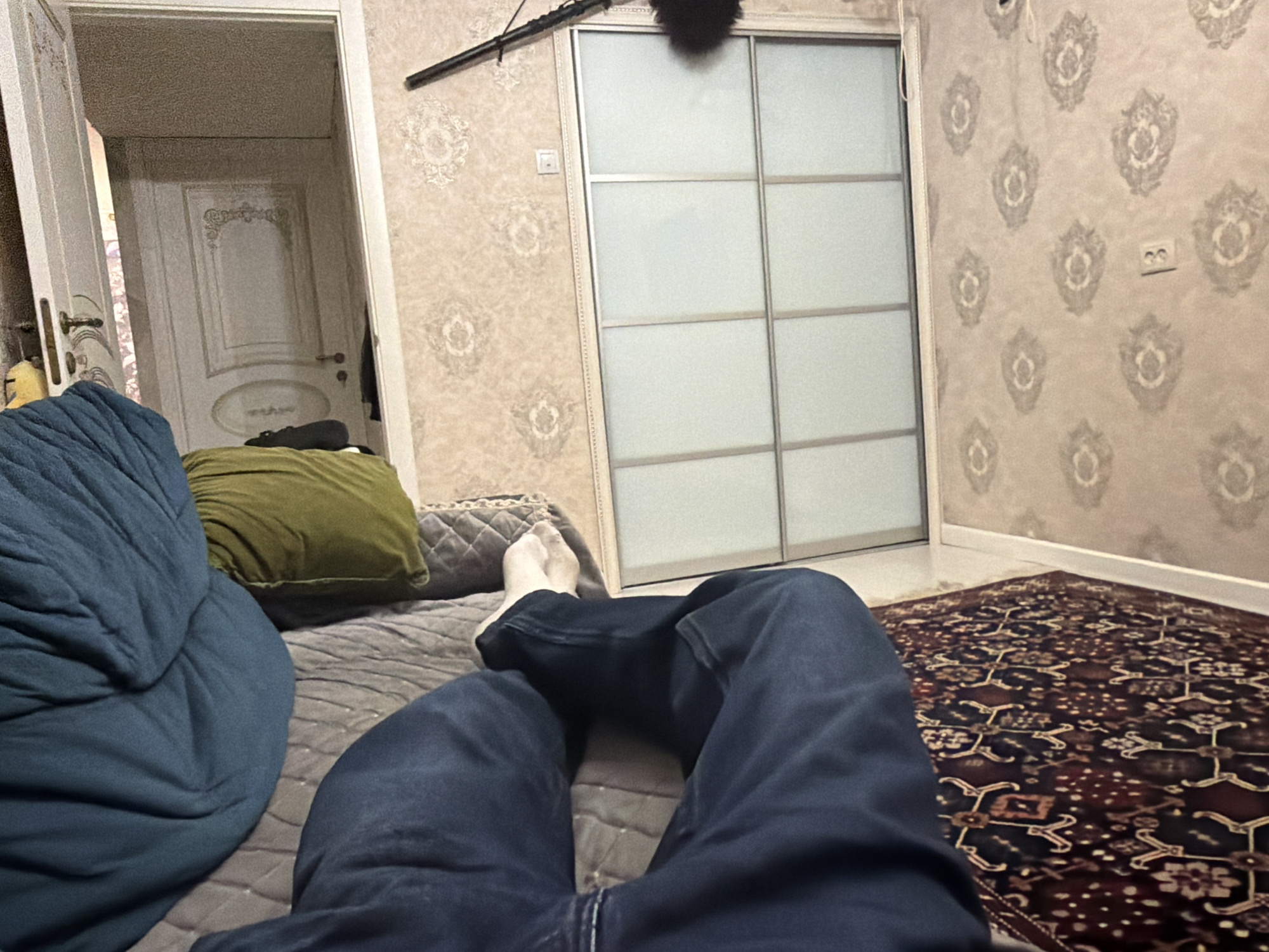 The image depicts a first-person perspective of a person lying on a bed in a room. The person is wearing dark blue jeans and white socks. Their legs are bent, and their feet are visible in the foreground. To the left of the person's legs, a large, teal-colored blanket or comforter is draped over the bed. Behind this, a green pillow is visible. The bed appears to be covered with a grey quilted surface.
In the background, a sliding closet door with frosted glass panels and a silver frame occupies the right side of the image. To the left of the closet, a doorway leads to another room. This doorway reveals a white-paneled door with decorative molding and a silver handle.
The walls are covered in a textured wallpaper with a repeating damask-like pattern in shades of beige and grey. A dark-colored object, possibly a camera or microphone, is mounted on the wall above the closet. Electrical outlets are visible on the wall. A dark, ornate rug with red and black patterns lies on the floor in front of the closet. The lighting in the room appears to be artificial, casting a warm glow. The overall impression is of a bedroom or living space. The location is specified as Unknown, Russia.