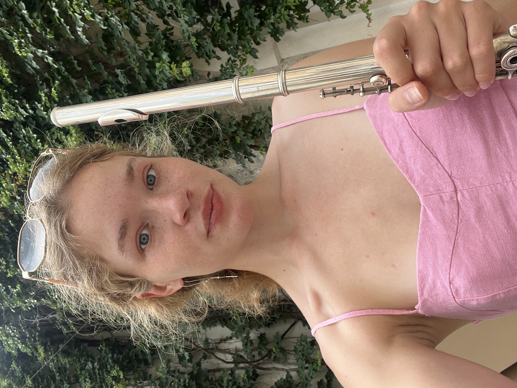 The image depicts a young woman with light skin and blonde hair, wearing a pink sleeveless top. She is holding a silver flute. Sunglasses are perched on her head. The background features green foliage. It is not possible to ascertain the specific location in Budva, Montenegro from this image alone.