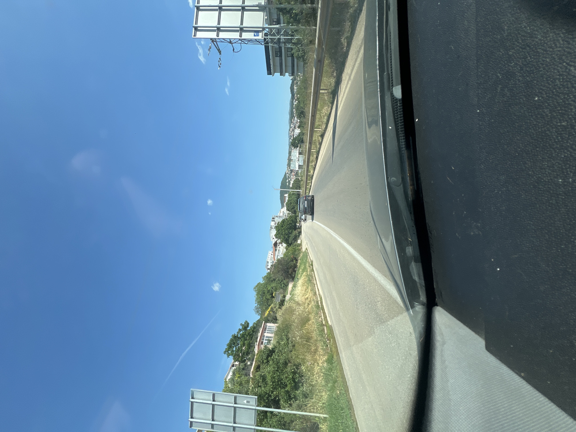 This is a first-person perspective, vertical video taken from inside a vehicle. The viewer is looking out of the passenger side window onto a road that curves away into the distance. The sky is a vibrant, clear blue with a few wispy white clouds. On the right side of the frame, the dark interior of the vehicle is visible, including part of the dashboard.
The road is asphalt with a white dividing line. There is a guardrail on the left side of the road, separating it from a grassy embankment. Beyond the embankment, there are trees and buildings, suggesting a town or village is situated on a hillside. The architecture appears to be white buildings with red-tiled roofs, consistent with Mediterranean or Balkan styles. A dark-colored car is visible driving ahead on the road.
On the left side of the road, further up the embankment, there are more buildings and lush green vegetation. A large, rectangular billboard or sign structure stands prominently near the edge of the road in the distance. The sun appears to be high in the sky, indicating it is daytime, likely late morning or early afternoon, given the bright light and clear conditions. The overall scene suggests a sunny day in a coastal or hilly region.
The location is identified as Dobra Voda, Montenegro, which is a coastal town. This context aligns with the visual cues of a sunny climate, vegetation, and buildings on a hillside overlooking what is likely the Adriatic Sea (though the sea itself is not directly visible). No people are clearly discernible in the video. The activity is simply driving through this scenic area.