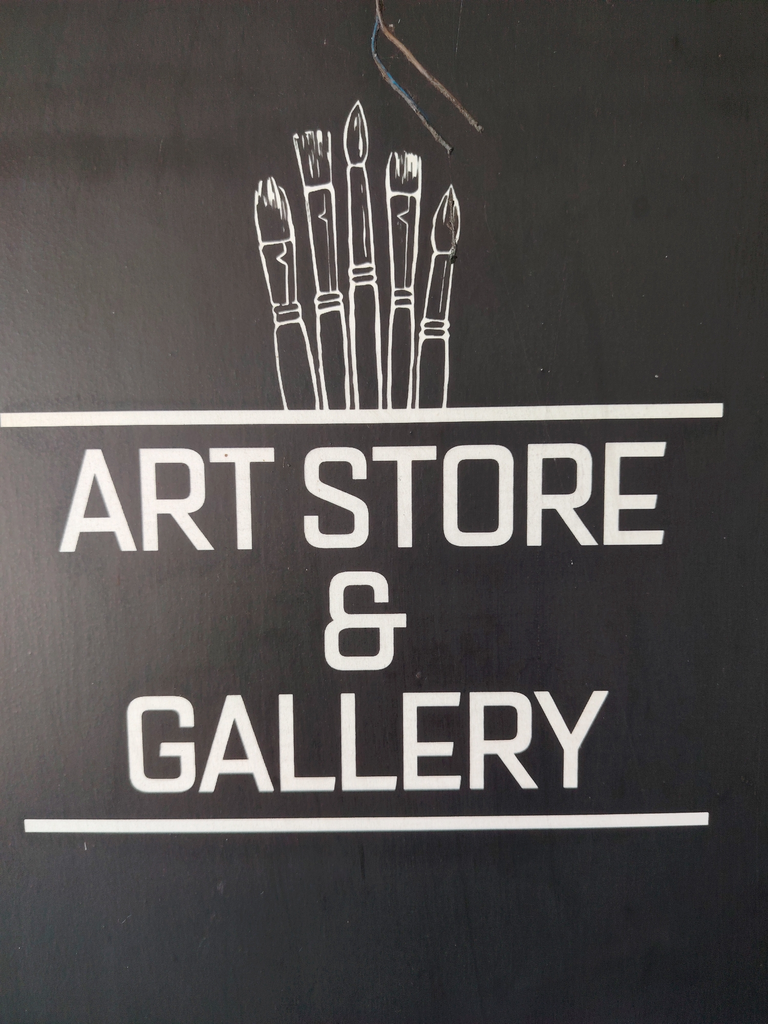 A black sign with white lettering displays the words "ART STORE & GALLERY." Above the text, a line of white paint brushes is depicted. The sign is mounted on a wall or a similar flat surface, with some discolored wires visible at the top of the image. The overall context is that of a storefront or commercial establishment.