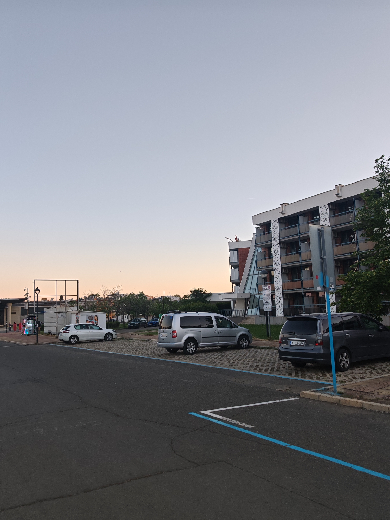 The image shows an outdoor parking area adjacent to a multi-story building, possibly a hotel, during what appears to be early evening or dusk. The sky is a gradient of light blue to orange near the horizon.
In the parking area, there are three cars visible: a white hatchback, a silver minivan, and a dark gray SUV. The SUV, parked in the foreground, has a visible license plate "A 2580 PP". The parking spaces are marked with blue lines.
To the left, a structure with a billboard frame is visible, along with what seems to be a small fairground attraction (likely near an open-air market or promenade) . Some people, dressed in light-colored clothing, are standing in front of it.
The building on the right side of the image is a multi-story structure with balconies. The architecture features white horizontal lines and dark ornamental lattice on the balconies.
This location is likely in or near Nesebar, Bulgaria, based on the Bulgarian license plate and the architectural style, typically seen in tourist areas of the Bulgarian Black Sea coast.