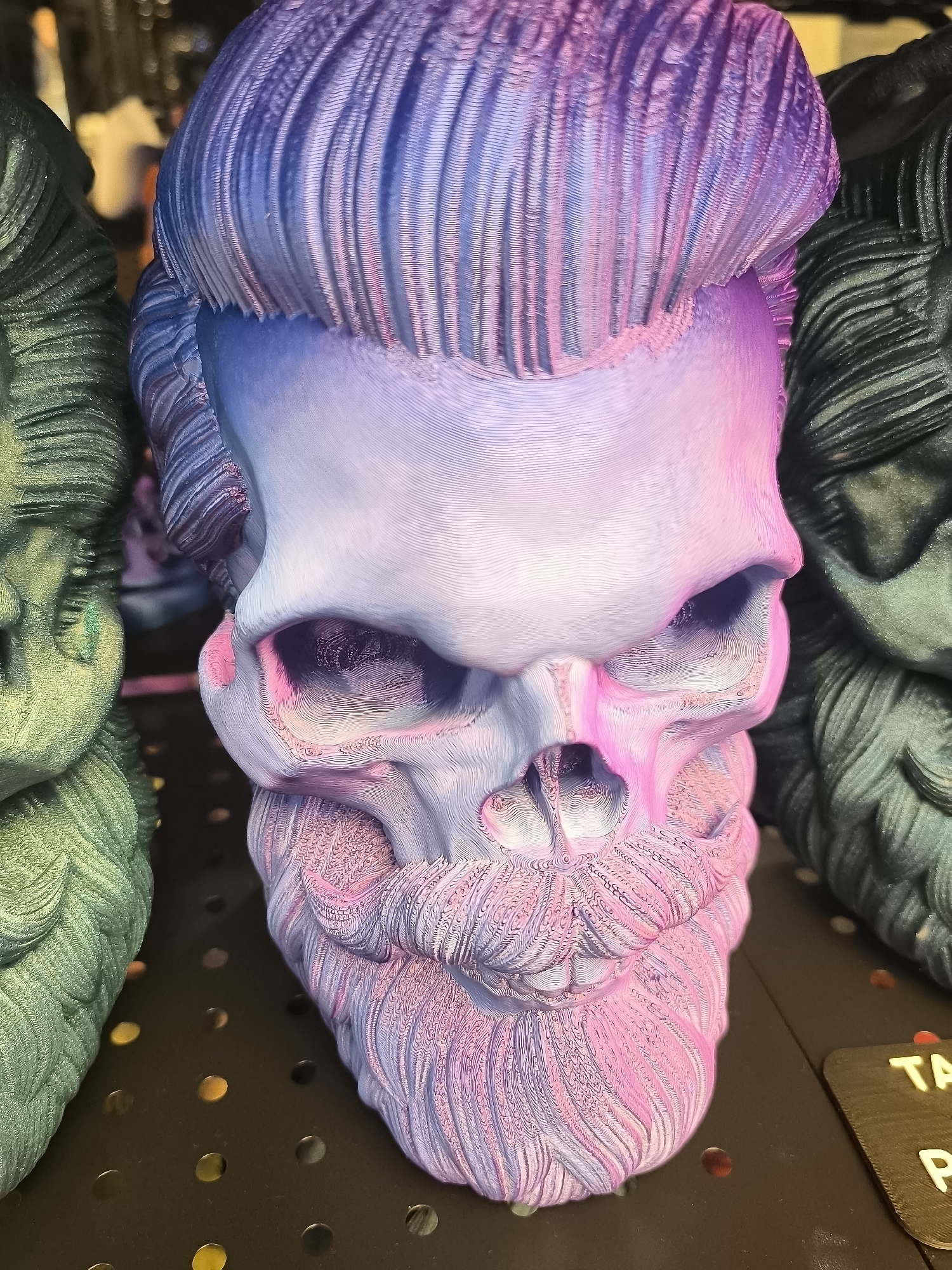 This is a close-up, eye-level shot of a 3D-printed skull with a stylized beard and hair. The skull is colored with a gradient of purple, pink, and light blue, with the hair appearing more intensely purple and the beard more pink. The skull has prominent eye sockets and a textured, ribbed surface. It is positioned centrally in the frame, with parts of other similarly textured 3D-printed objects, one appearing green and the other dark, on either side. The background is dark and out of focus, with some blurry lights visible. A small, dark brown tag with the letters "TA" and "P" in yellow is visible in the lower right corner. The scene is indoors, likely a retail or exhibition space, possibly a shop selling 3D printed items. There is no identifiable information to place this image in Klaipėda, Lithuania.