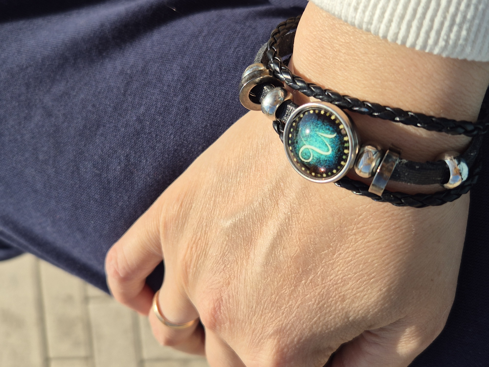 A close-up shows a person's hand wearing a multi-strand black braided leather bracelet with silver-tone metal embellishments. A circular pendant on the bracelet features a teal and blue zodiac sign with a starry background. The person wears a simple gold ring on their ring finger. Their hand is resting on a navy blue fabric, possibly clothing. The background shows a paved surface suggesting an outdoor setting. The image's sharp focus is on the hand and bracelet, with the surroundings slightly blurred. There is no specific indication of this being in Kokshetau, Kazakhstan.