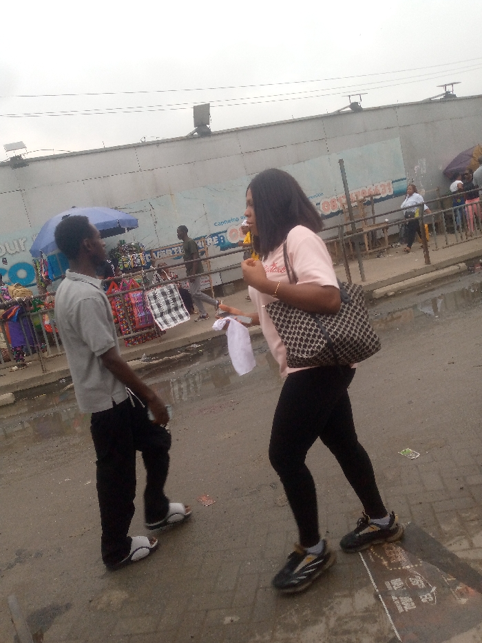 The image depicts a street scene in Badore, Nigeria, under an overcast sky. In the foreground, two individuals are walking on a paved surface. A female, positioned on the right, wears a light pink t-shirt, black leggings, and black athletic shoes with yellow accents. She carries a black and white patterned shoulder bag on her left shoulder and holds a white cloth or paper in her left hand. Her dark hair is shoulder-length. Opposite her, on the left, a male wears a light grey polo shirt, black trousers, and white slide sandals. Both individuals are looking towards each other.
In the mid-ground, behind the subjects, a long, grey corrugated metal or panel wall extends horizontally. Faded blue text is visible on the wall, including "Capturing" and a partial phone number "081...". Above the wall, utility lines stretch across the frame, and dark rectangular structures, possibly vents or small solar panels, are visible on the roofline. To the left, under a blue umbrella, market stalls display various goods, including brightly colored fabrics and black and white checkered bags. Several other individuals are visible in the background, walking or standing near the stalls. The ground appears damp with some puddles and scattered debris.