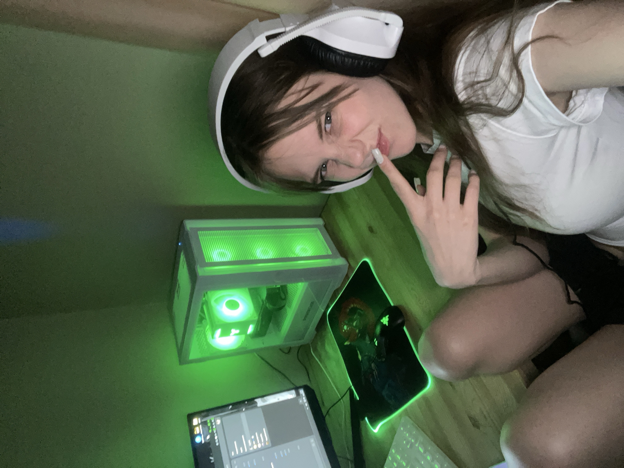 A young woman with brown hair, wearing a white t-shirt and black shorts, sits in a room. She is also wearing white headphones. She holds a finger to her lips. A computer tower with green LED lighting sits on a desk next to a monitor and a mousepad with green LED edging. The image is dimly lit and the environment appears to be indoors, against a green wall. No specifics indicate this photo was taken in Ufa, Russia.