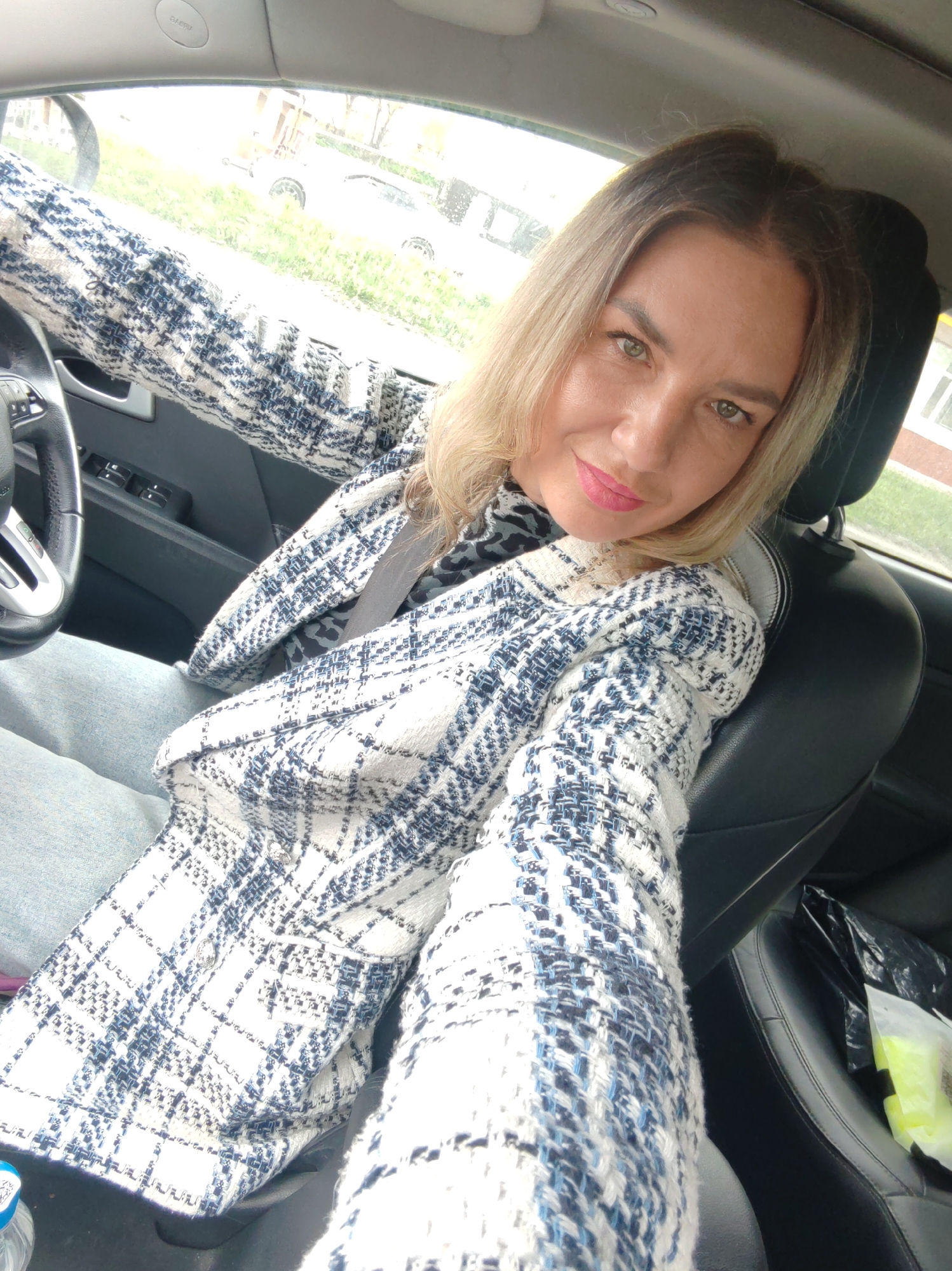 A woman with blonde hair and green eyes is taking a selfie inside a car. She is wearing a white and blue patterned jacket and a gray seatbelt is visible. The steering wheel and car door are also visible. Outside the car, there is grass and buildings. The image appears to be taken in the city of Petropavlovsk-Kamchatsky, Russia.