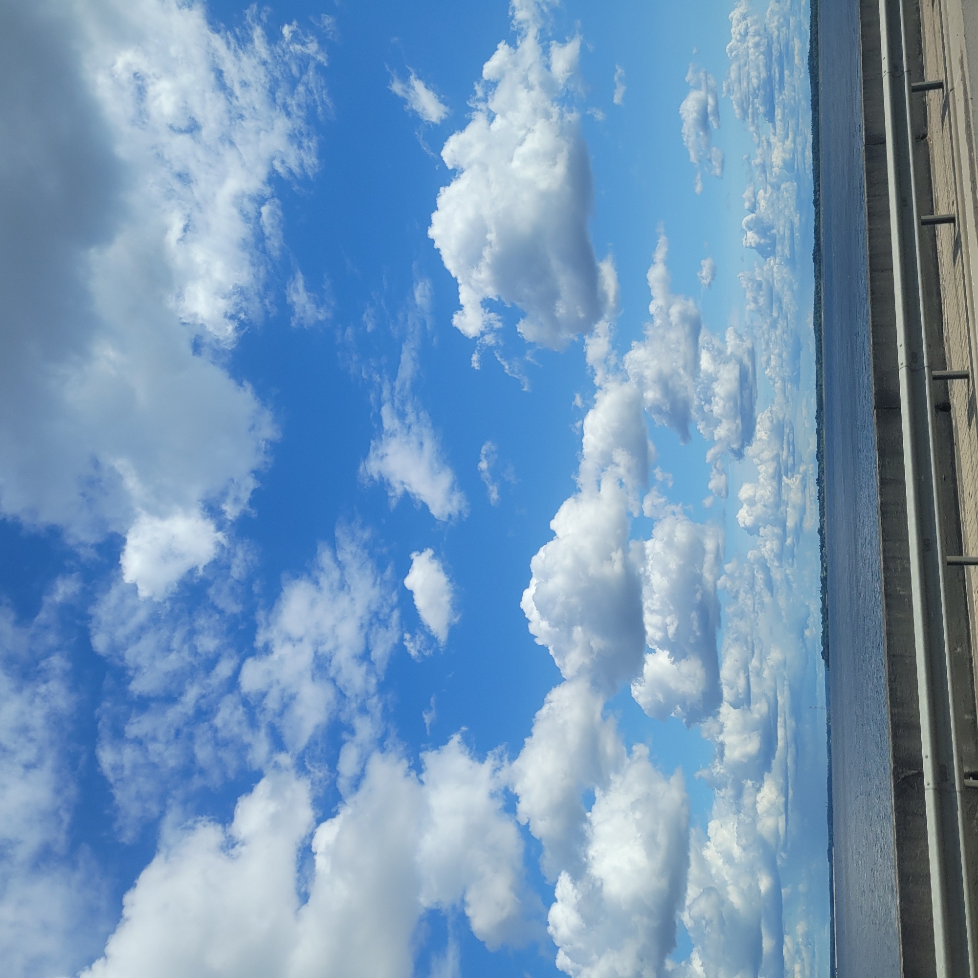 The image captures a vast expanse of the sky filled with scattered white and gray cumulus clouds against a bright blue backdrop. Below the sky, a wide, dark blue body of water stretches horizontally across the frame, with a faint, tree-lined horizon visible in the distance. In the foreground, a concrete barrier with a metallic railing runs along the bottom of the image, suggesting a bridge or elevated walkway overlooking the water. The scene is devoid of any people or explicit signs of activity, presenting a serene, natural landscape.