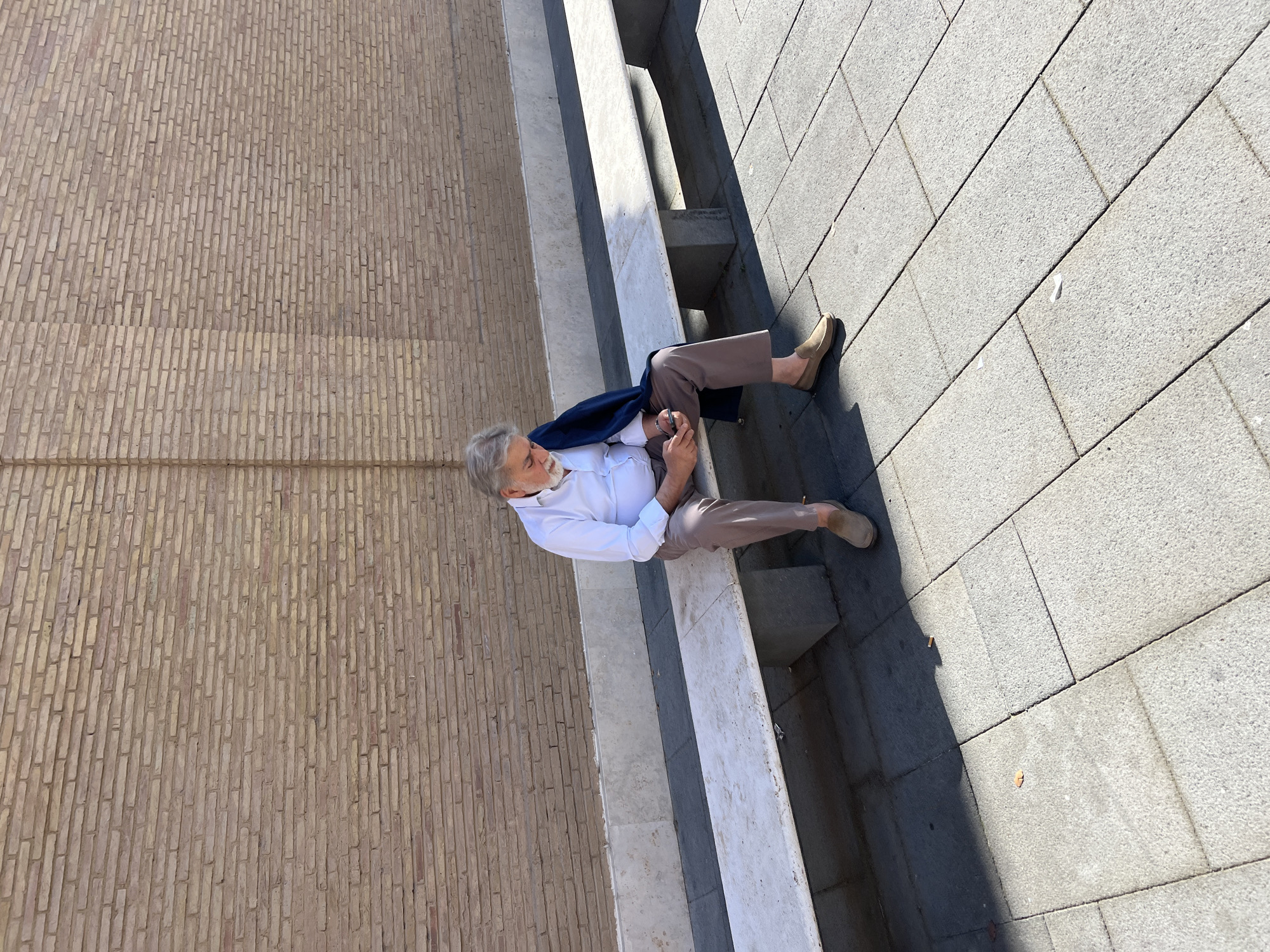 The scene depicts an elderly male seated on a stone bench outdoors, under bright daylight, in an urban setting. The location is consistent with the external walls of the Vatican City in Rome, Italy, possibly along Viale Vaticano.
The male, with grey hair and a full grey beard, wears a white long-sleeved shirt, light brown trousers, and beige slip-on loafers. A dark blue jacket is draped over his left shoulder. He is looking down at a smartphone held in both hands. He is seated on a long, light-colored stone bench with visible wear and tear, supported by darker, rough-hewn stone blocks.
Behind the bench stands a tall, weathered brick wall, predominantly light brown and yellowish in tone, constructed with horizontal stretcher bond courses. A vertical architectural element resembling a pilaster or recessed column is visible in the brickwork to the left of the man. A plinth of lighter, rough stone runs along the base of the brick wall, directly beneath the bench.
The ground is paved with rectangular grey stone slabs, characteristic of city sidewalks or plazas. Strong sunlight casts clear shadows on the ground and bench. Small pieces of refuse, including what appears to be a discarded cigarette butt, are visible on the paving stones near the bench. No other individuals are present, and there are no discernible interactions.